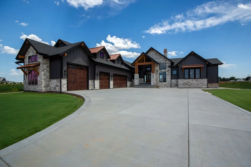 Modern house with stone and dark wood exterior, three garage doors, and a wide concrete driveway, under a partly cloudy sky.