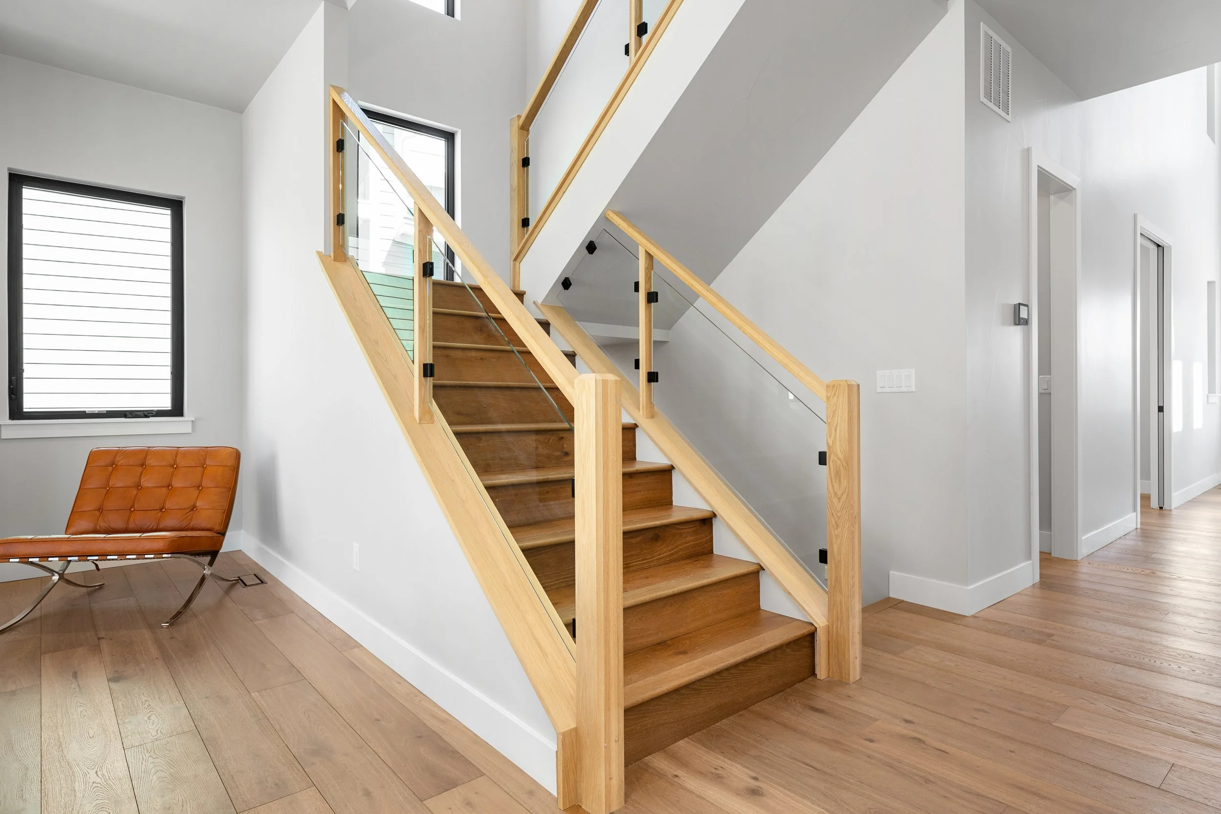 Interior of a modern home with wooden stairs, white walls, hardwood floors, and a leather chair, with windows and doors visible.