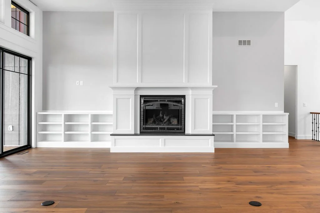 Empty living room with a white fireplace and built-in shelves on either side, hardwood floors, and a sliding glass door on the left.