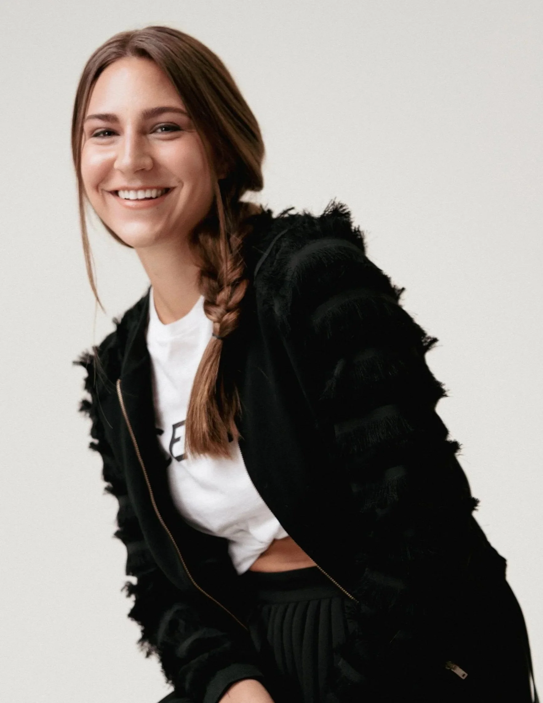 A smiling woman with long brown hair styled in a side braid, wearing a black textured jacket over a white shirt, standing against a plain white background.