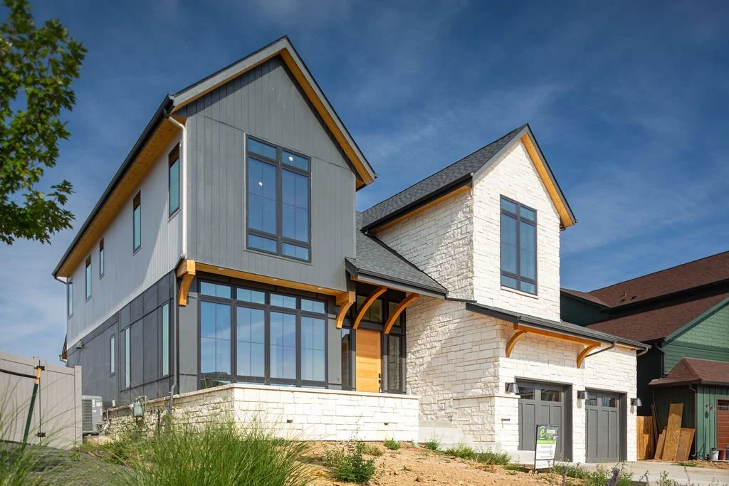 A modern two-story house with a mix of gray siding, white brick, and large windows under a blue sky.