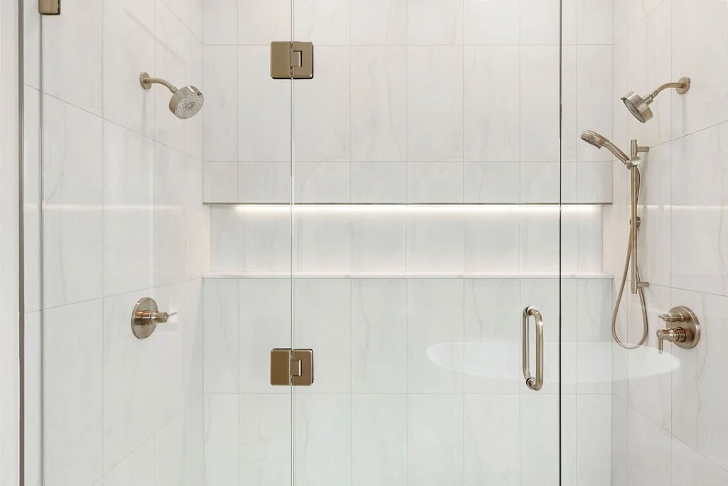 Inside a modern shower with a glass door, featuring two showerheads and a built-in shelf.