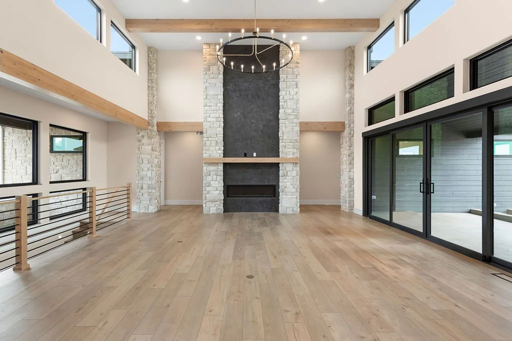 Modern living room with a high ceiling, large windows, a fireplace framed with white brick and black surround, a round chandelier, light wood floors, and sliding glass doors leading to an outdoor patio.