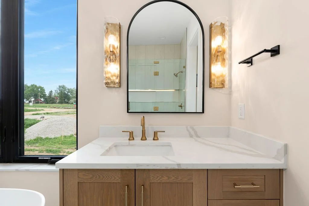 Bathroom vanity with a white marble top, a brass faucet, a wooden cabinet, a large arched mirror, two tall wall sconces with warm lighting, and a window showing an outdoor scene with green grass and trees.