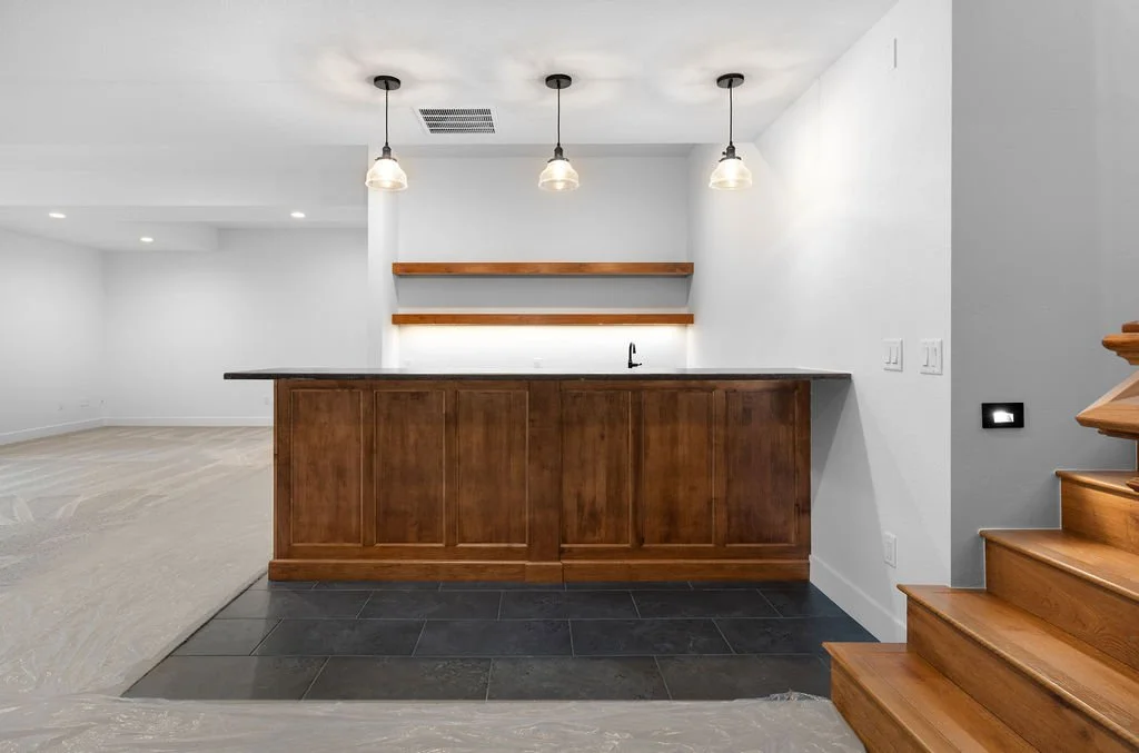 Empty kitchen with wooden bar, white walls, hanging pendant lights, and open modern living space in background.