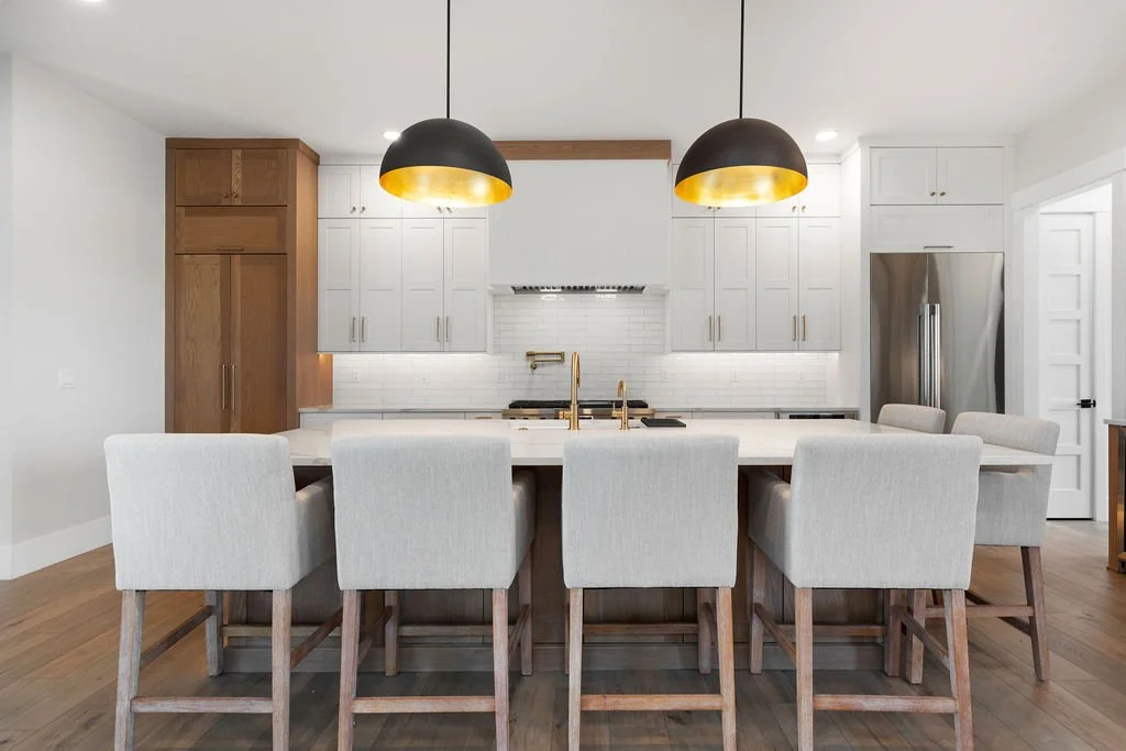 Modern kitchen with white cabinets, a large island, and eight beige chairs, with two black and gold pendant lights hanging overhead.