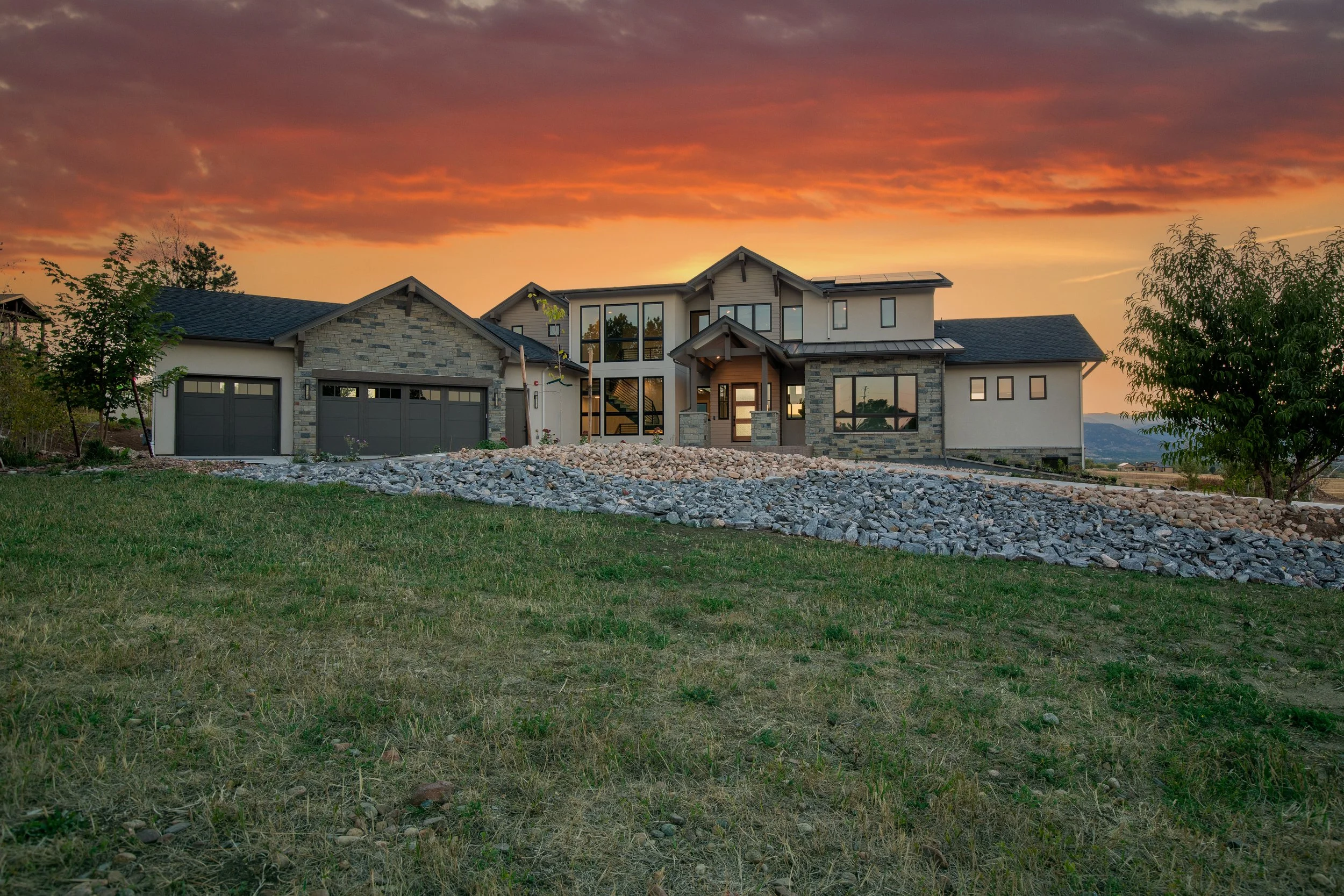Large modern house with a stone and siding exterior, multiple garage doors, and large windows, under a sunset sky with orange and pink clouds.
