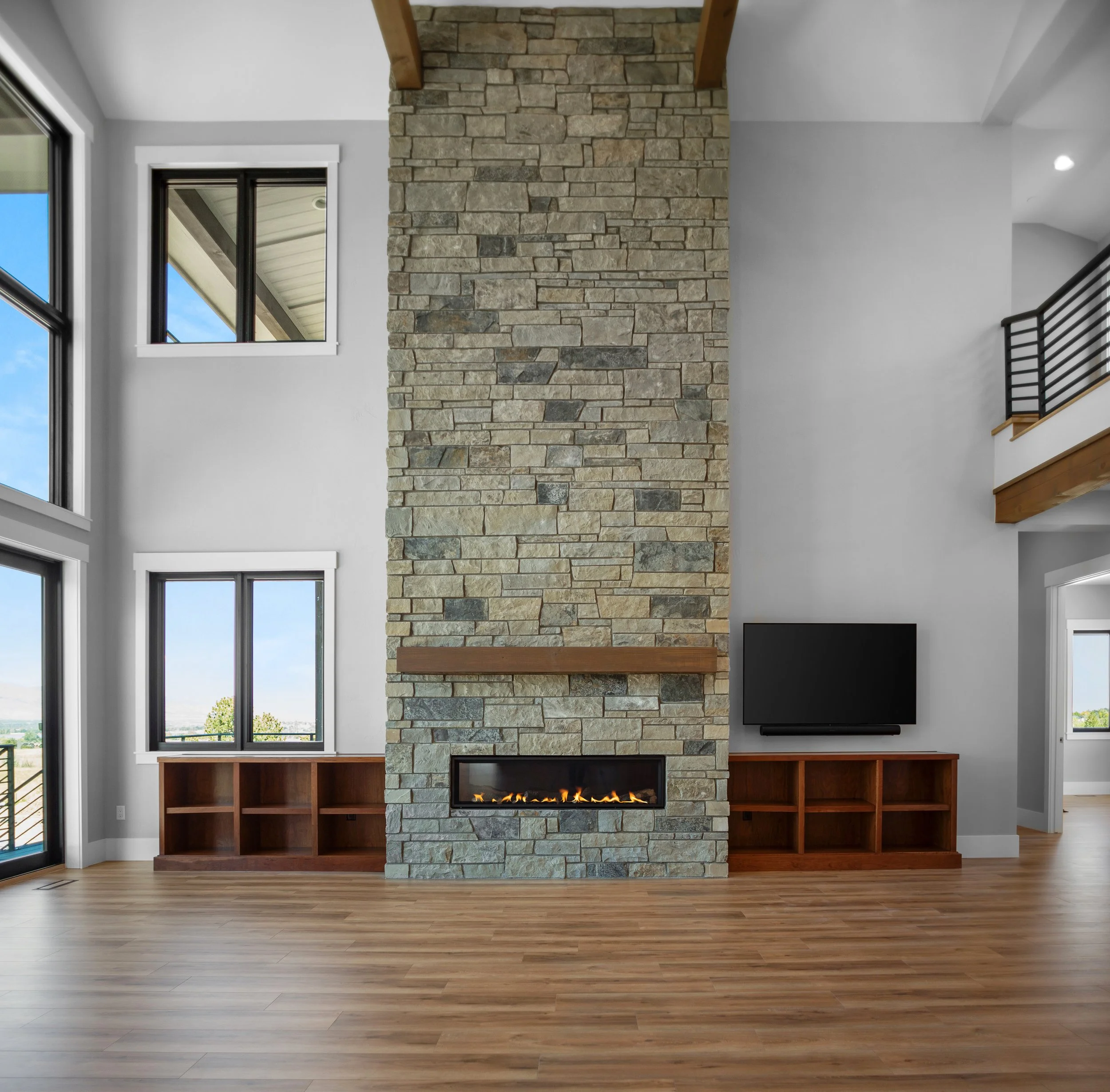Living room with a stone fireplace, wooden built-in shelves, a wall-mounted TV, and large windows with a view outside.