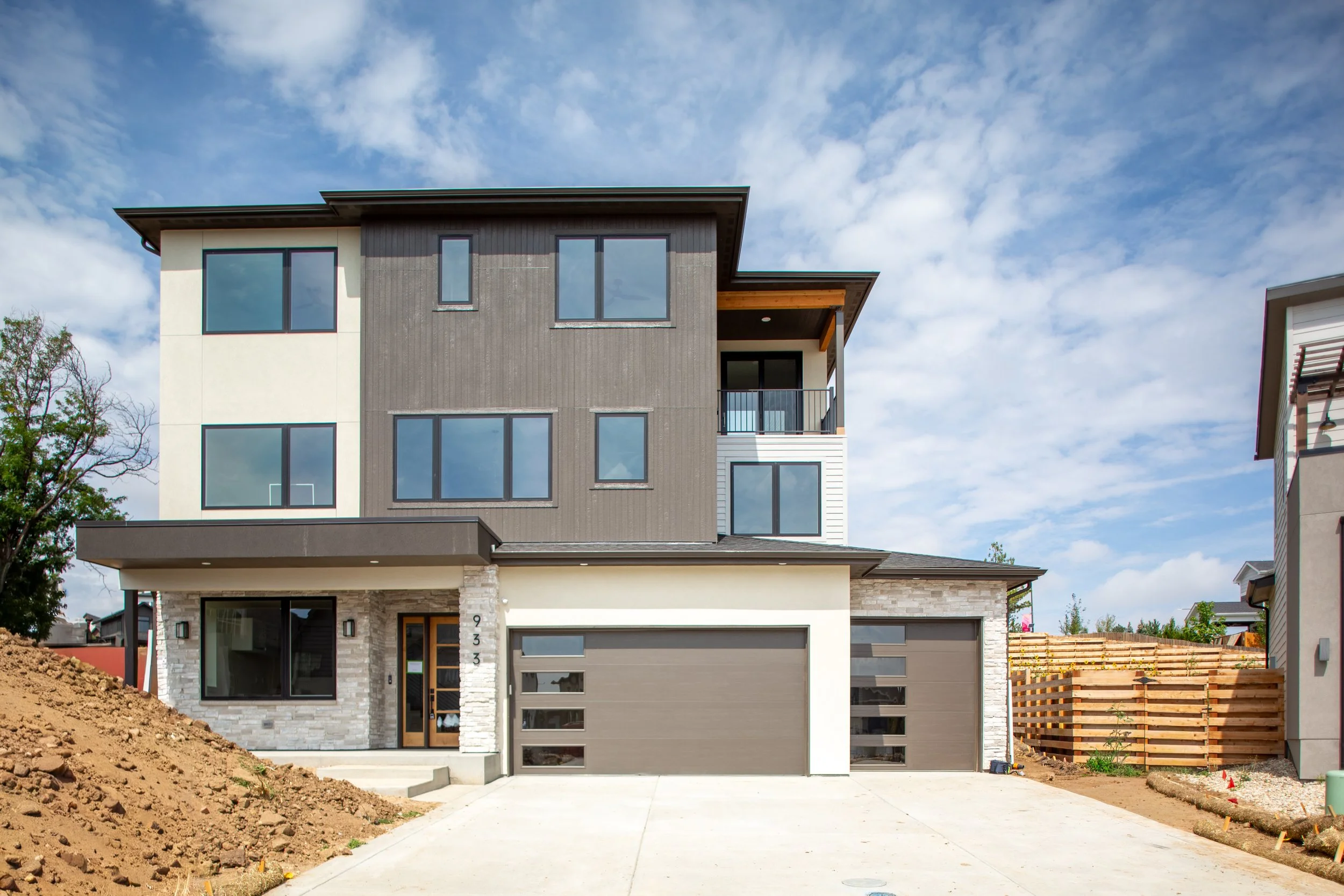 Newly constructed modern three-story house with a gray and white exterior, two-car garage, and a small front yard with dirt and paving.