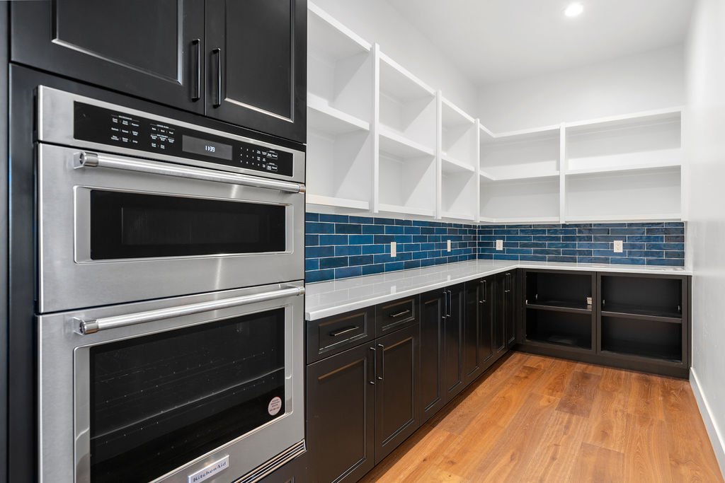 Kitchen with dark cabinets, white open shelving, blue subway tile backsplash, stainless steel double oven, white countertop, and wood flooring.