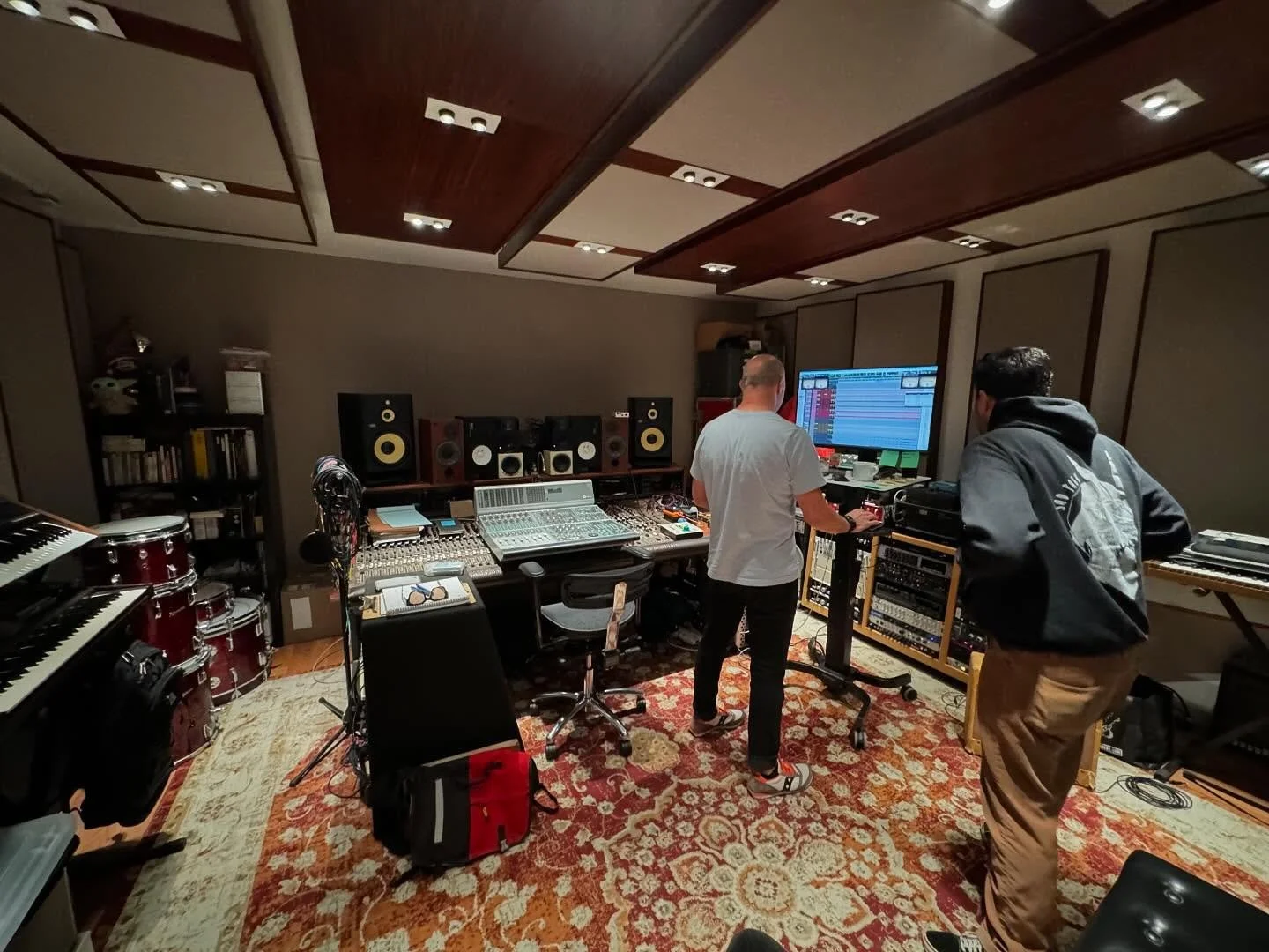 Two people working in a professional recording studio with various music equipment, speakers, and a large computer screen.