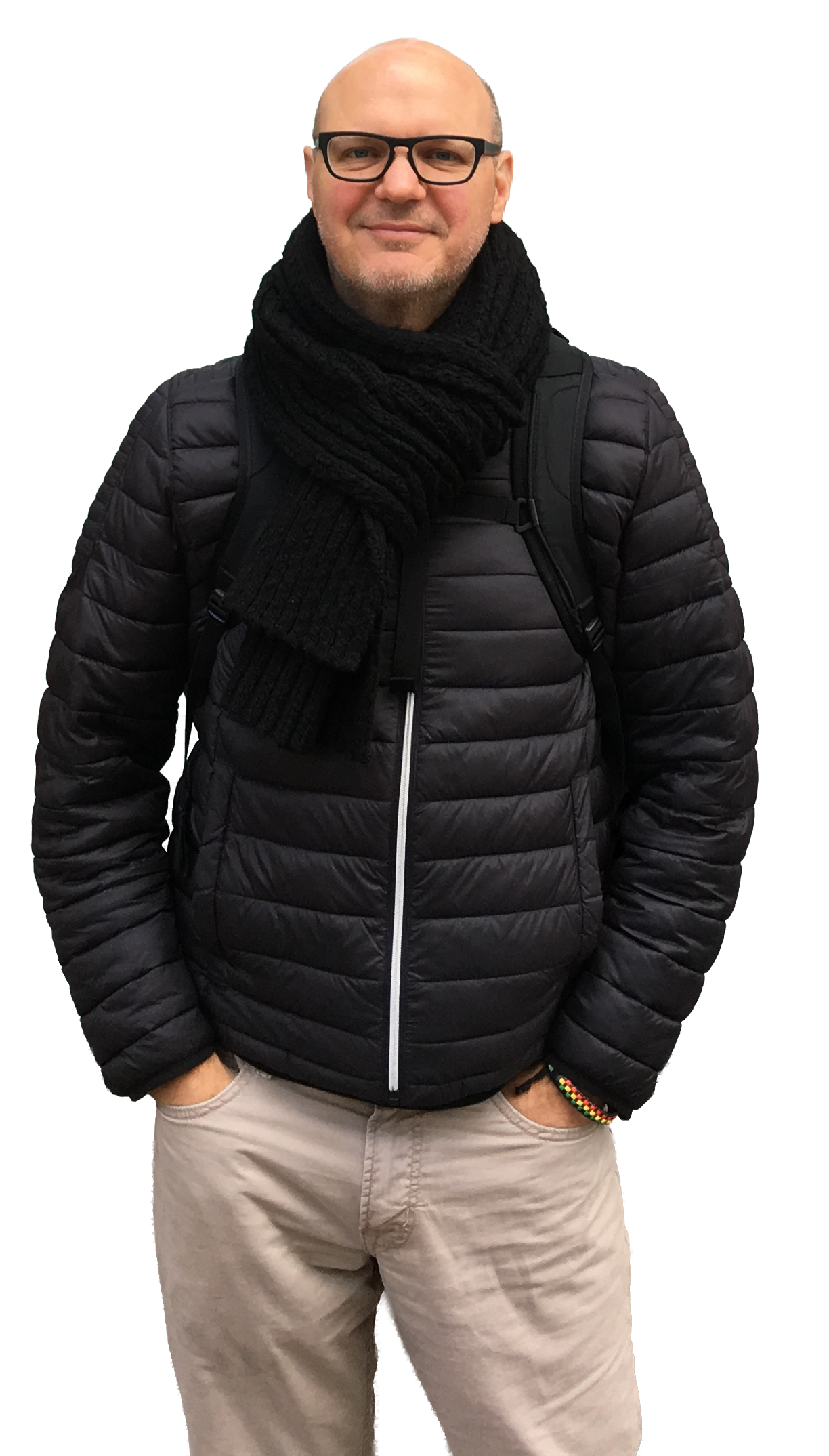 A man with glasses, dressed in a black puffer jacket, beige pants, and a black scarf, standing with hands in pockets and smiling.