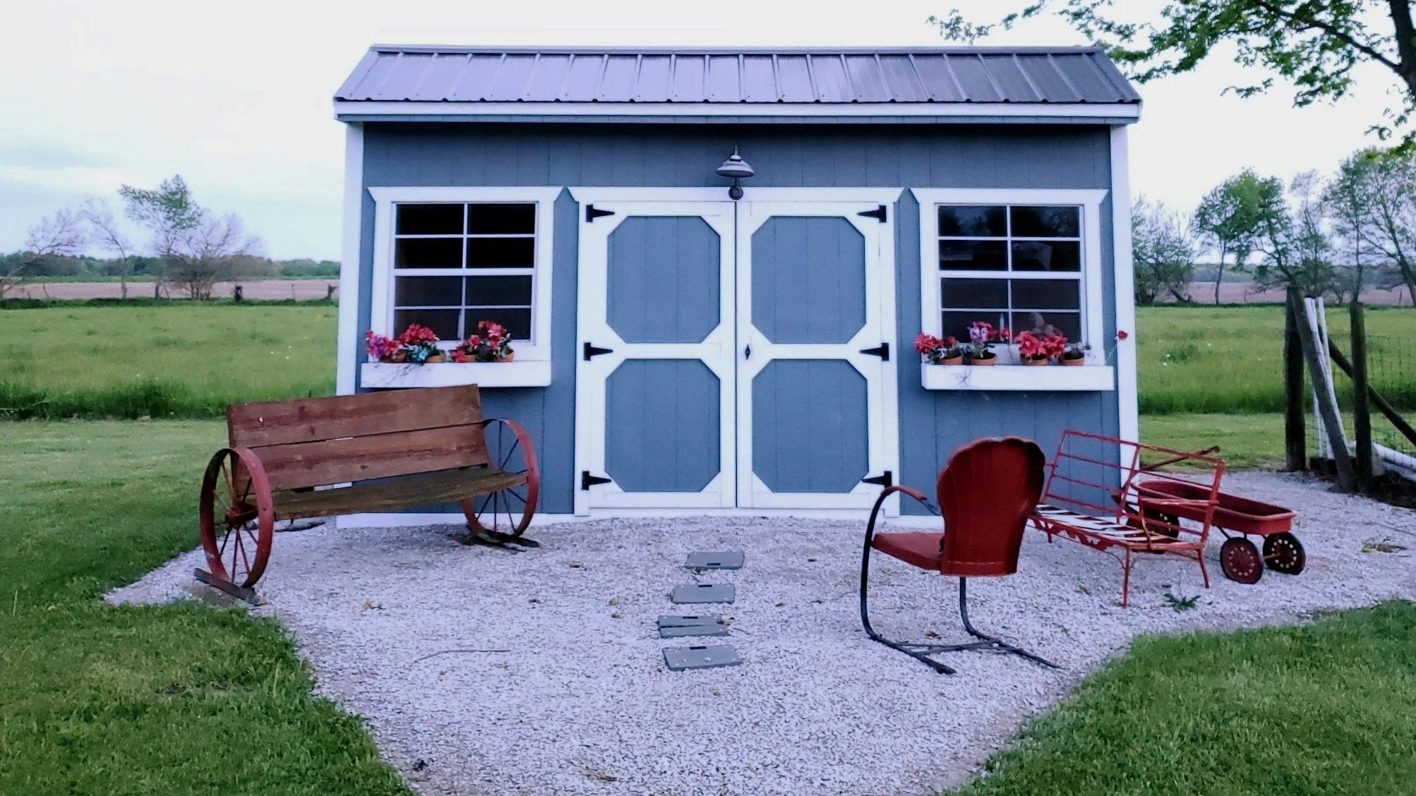 Blue garden shed with white trim, two window boxes with red flowers, and double doors, surrounded by green grass and trees. In front, there is a wooden bench with red metal wheels, a red metal chair, and a red wagon on gravel.