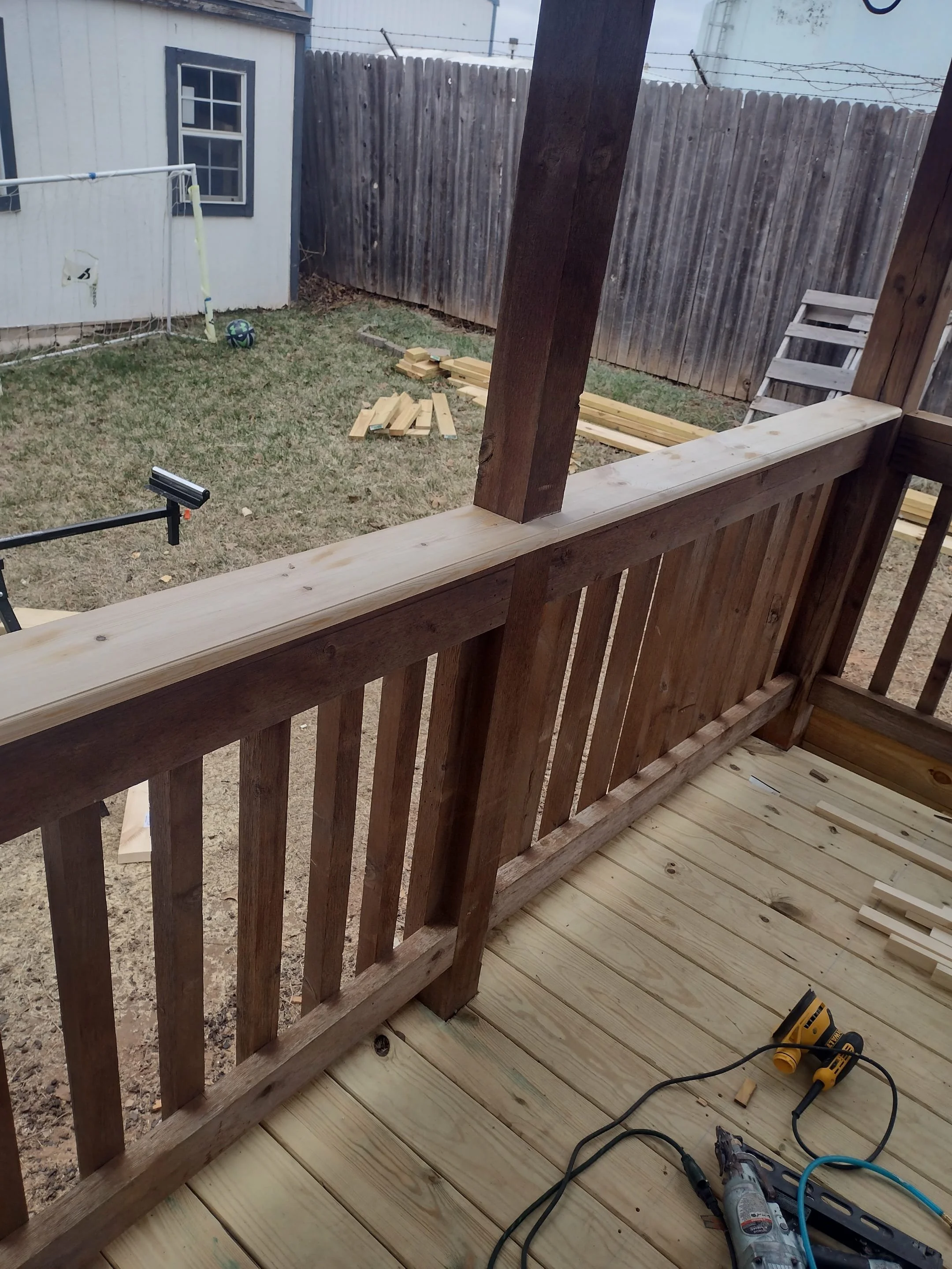 View of a backyard with a wooden deck under construction. Construction tools like a cordless drill, a measuring tape, and a saw are on the deck. The backyard has a small grassy area with scattered wood planks, a soccer ball, a small soccer goal, and 