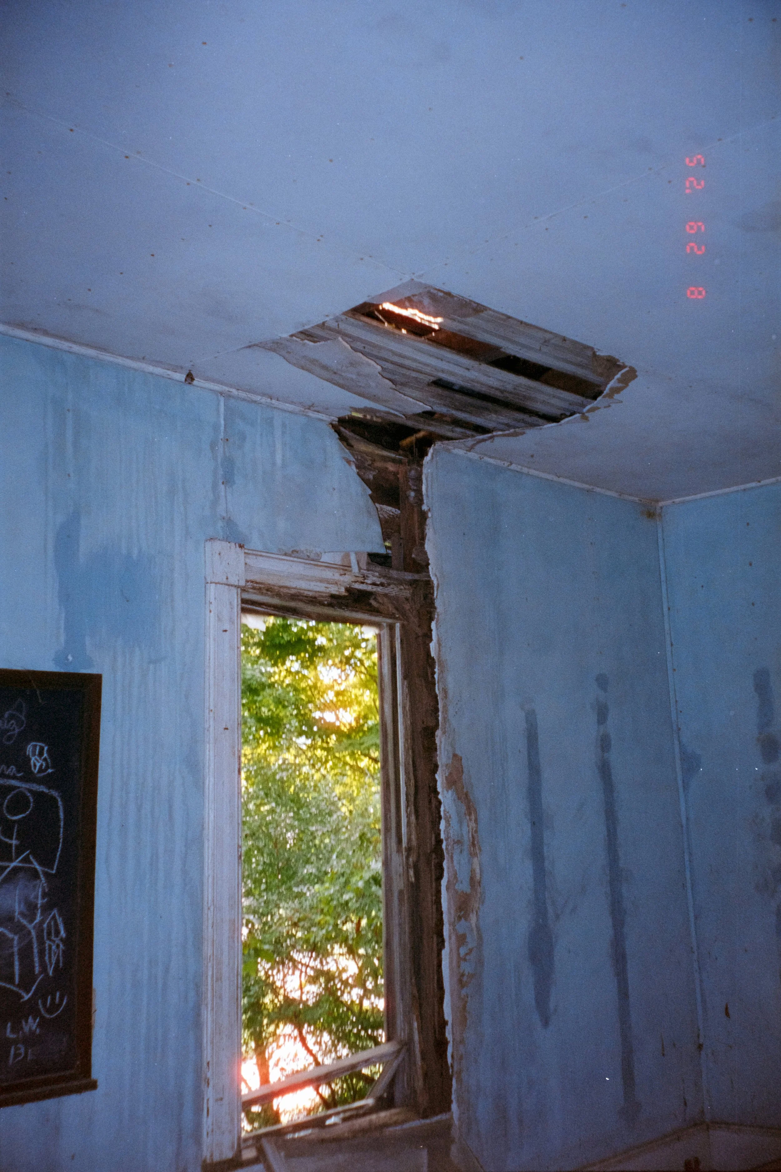 Interior of a damaged room with a hole in the ceiling and window frame, sunlight shining through a tree outside.