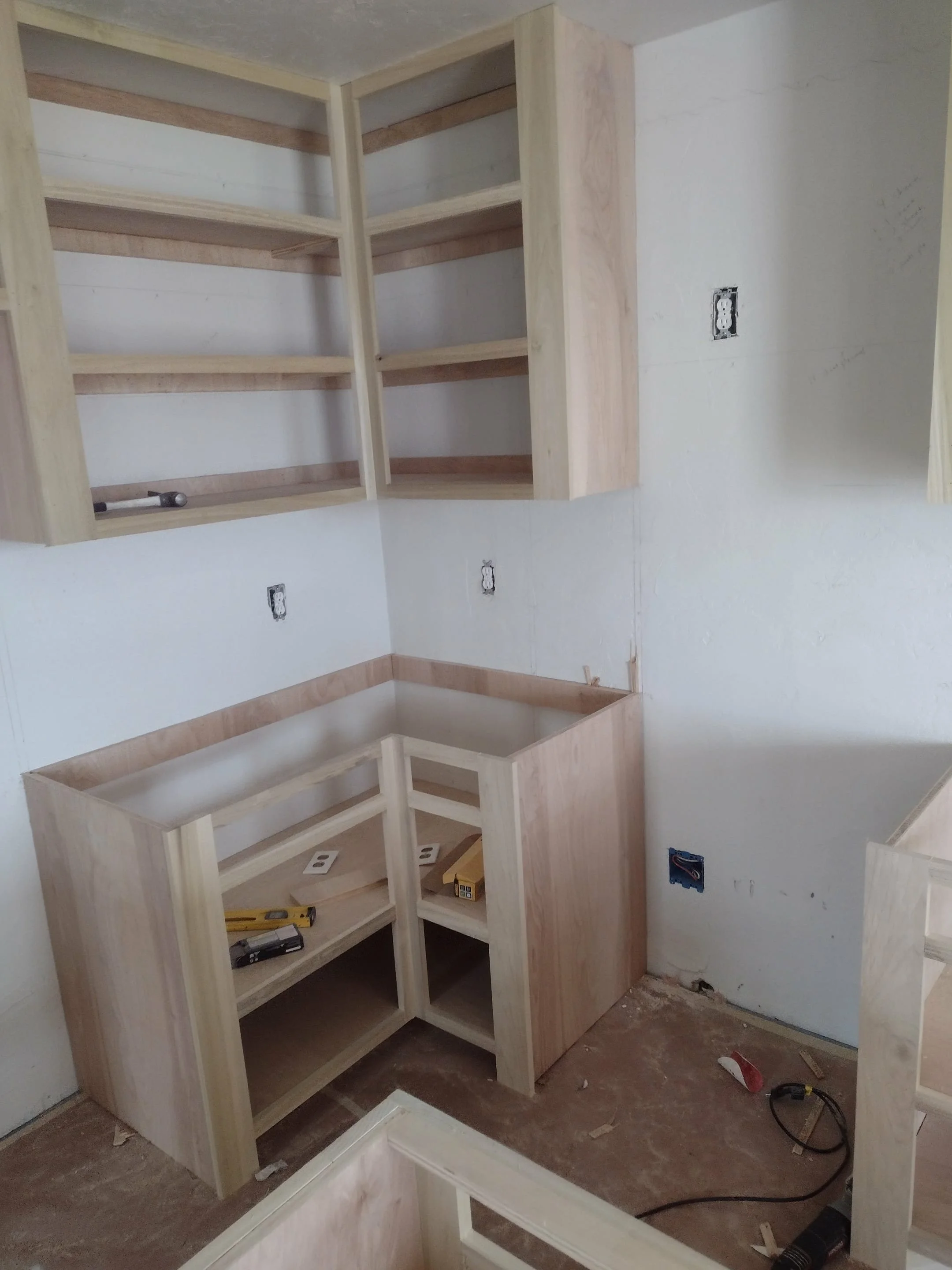unfinished kitchen cabinets being installed in a home under construction, with open shelving on the upper and lower sections, construction tools on the countertops, and electrical outlets on the walls.
