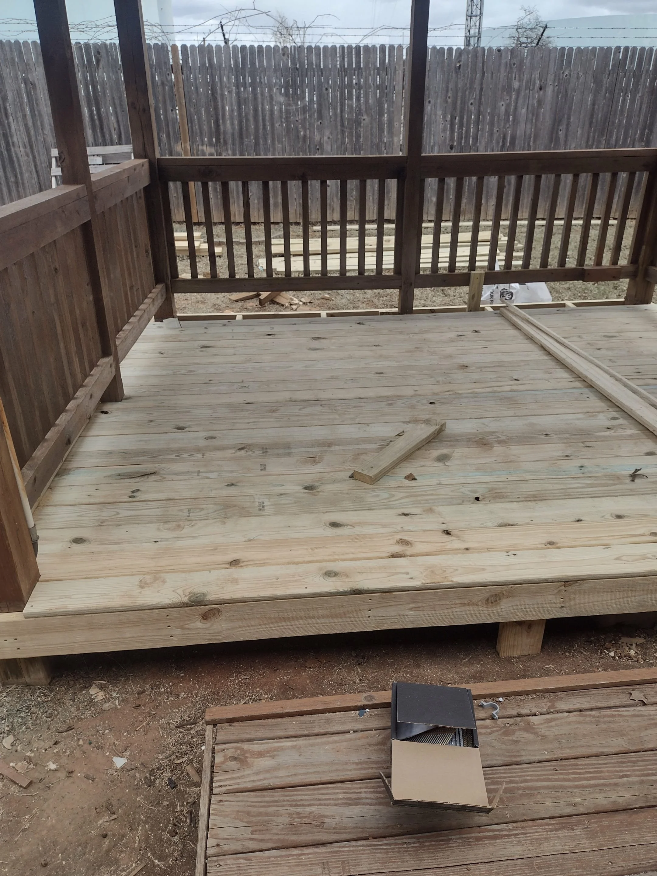 A newly built wooden deck under construction with wooden planks and posts, enclosed by a wooden fence, in a backyard with some construction materials and tools nearby.