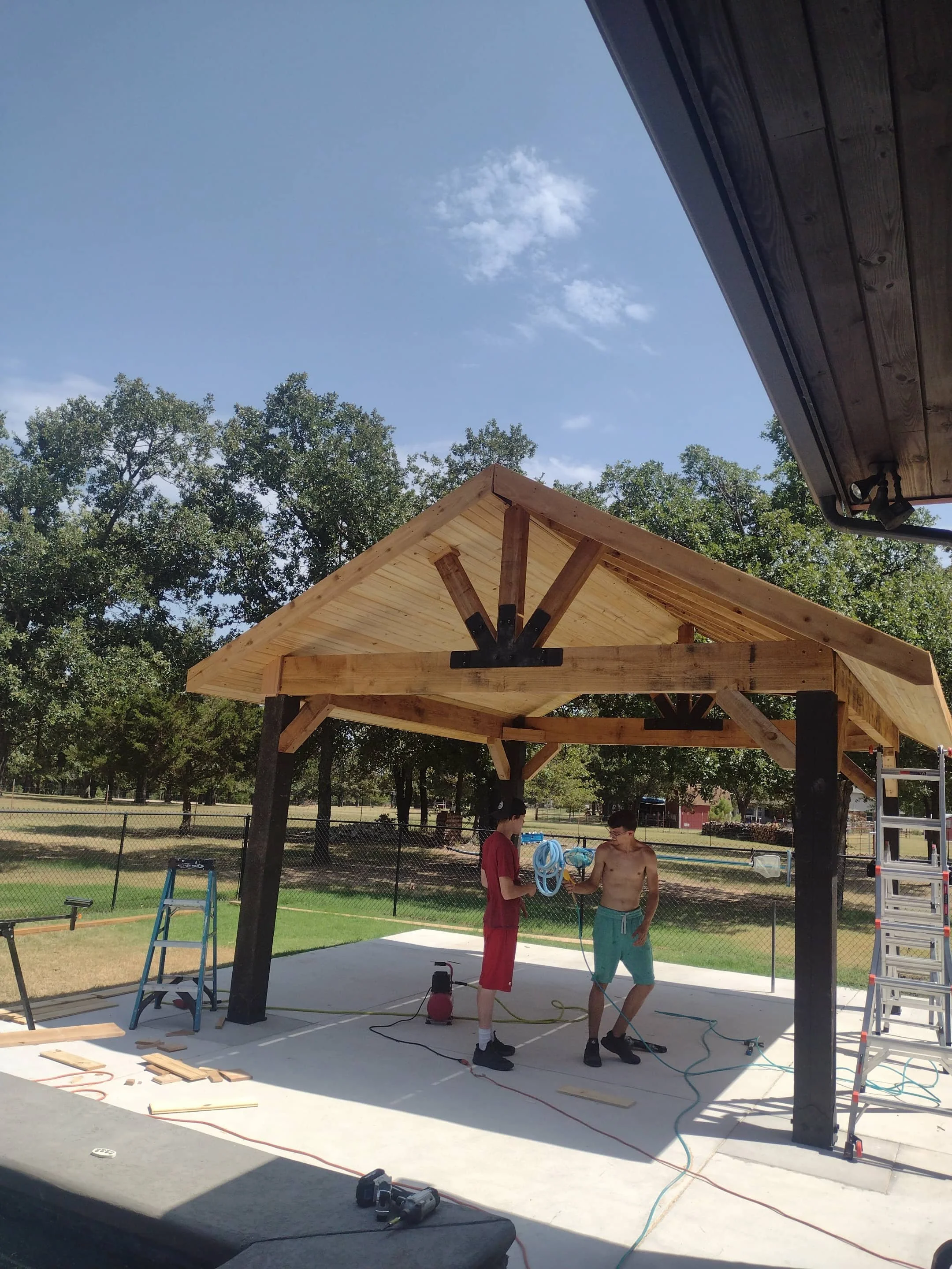 Two young men working on the construction of a wooden pavilion outdoors, with tools, ladders, and ropes around them, under a blue sky with some clouds.