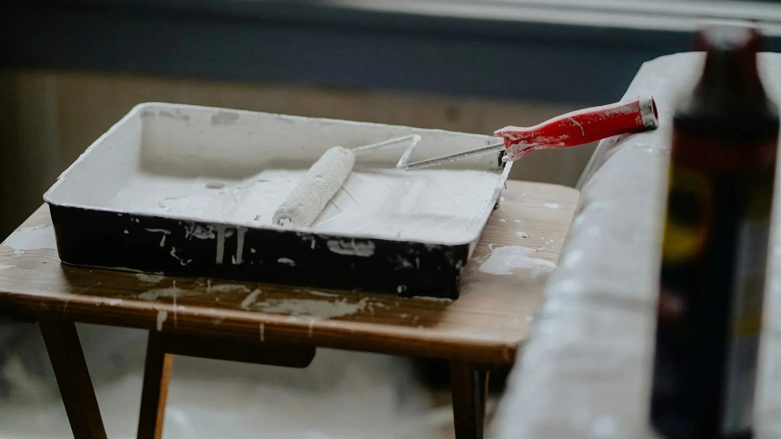 Paint roller, paintbrush, and container of white paint on a wooden table, with a roller tray, in a painting or renovation setting.
