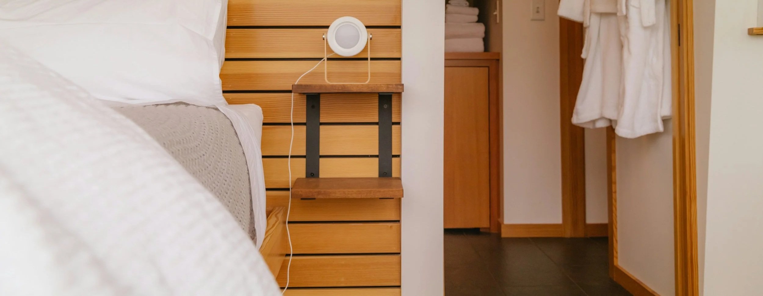 A bedroom with a bed, shoebox headboard, and a wooden nightstand with a white round speaker and charging cable, and a partially open closet with white bathrobes hanging inside.