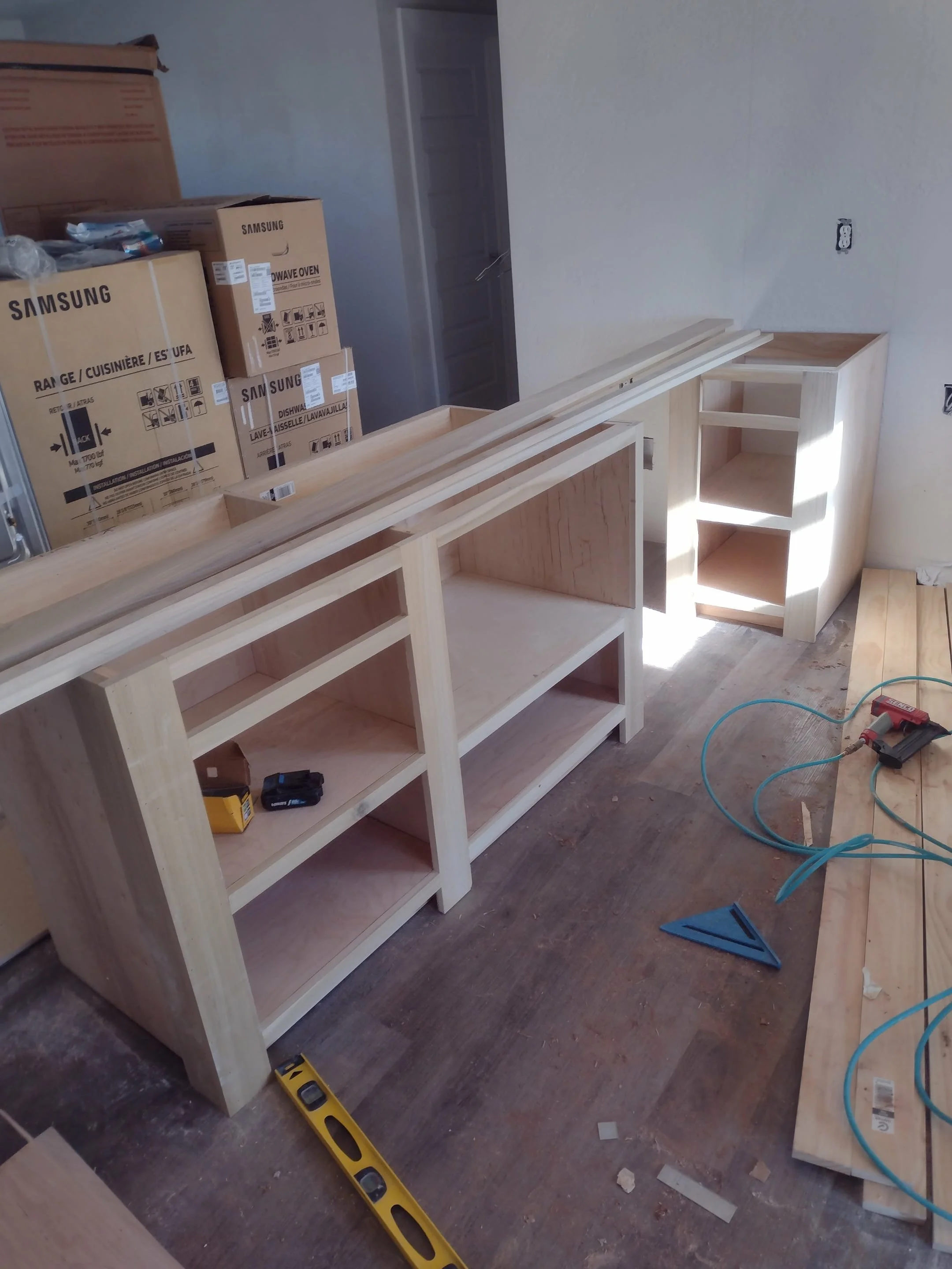 Wooden cabinets under construction in a room, with boxes of appliances in the background, construction tools and materials scattered on the floor.