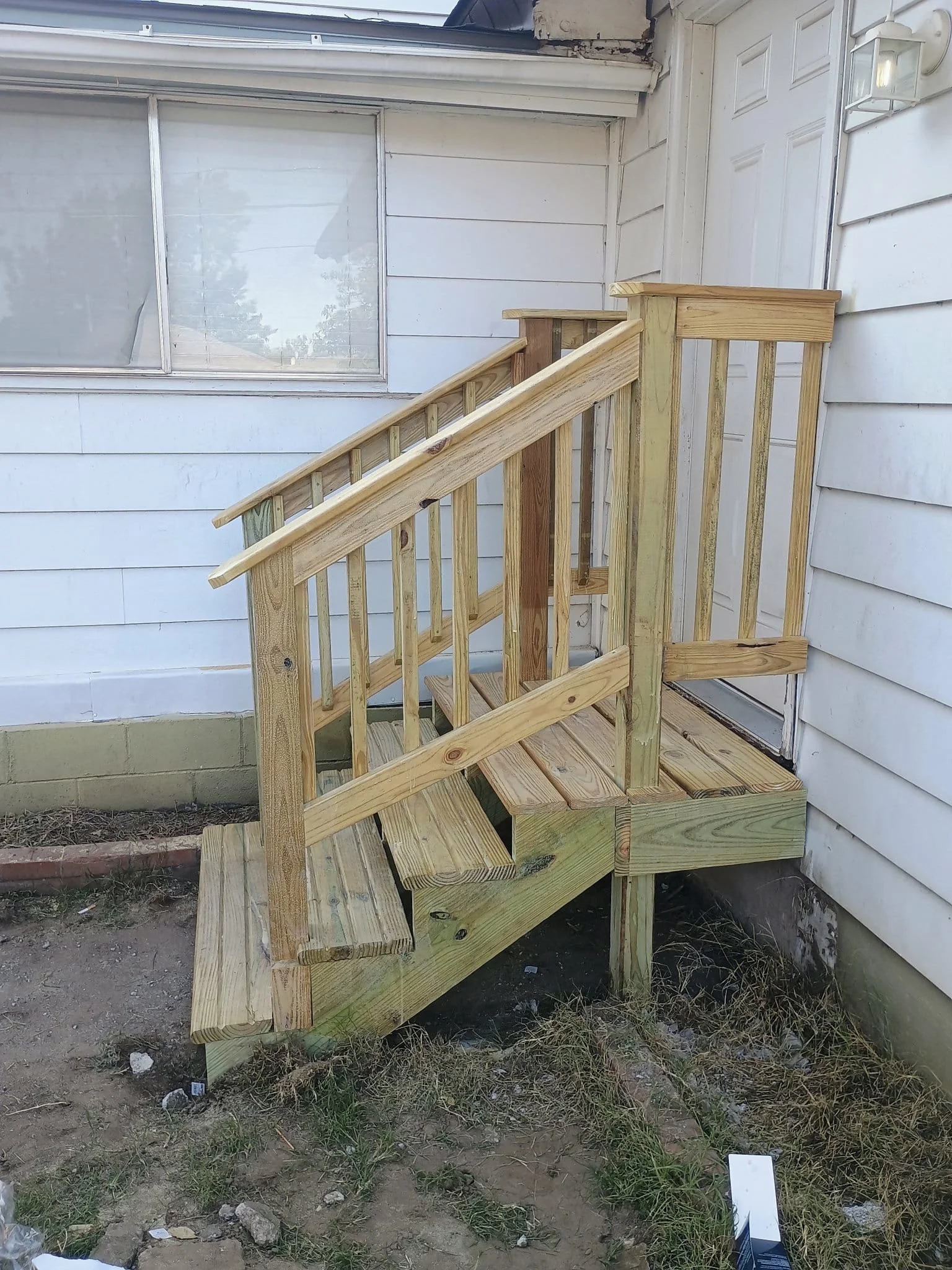 Newly built wooden staircase outside a house with white siding and a large window, partially installed with surrounding dirt and grass.