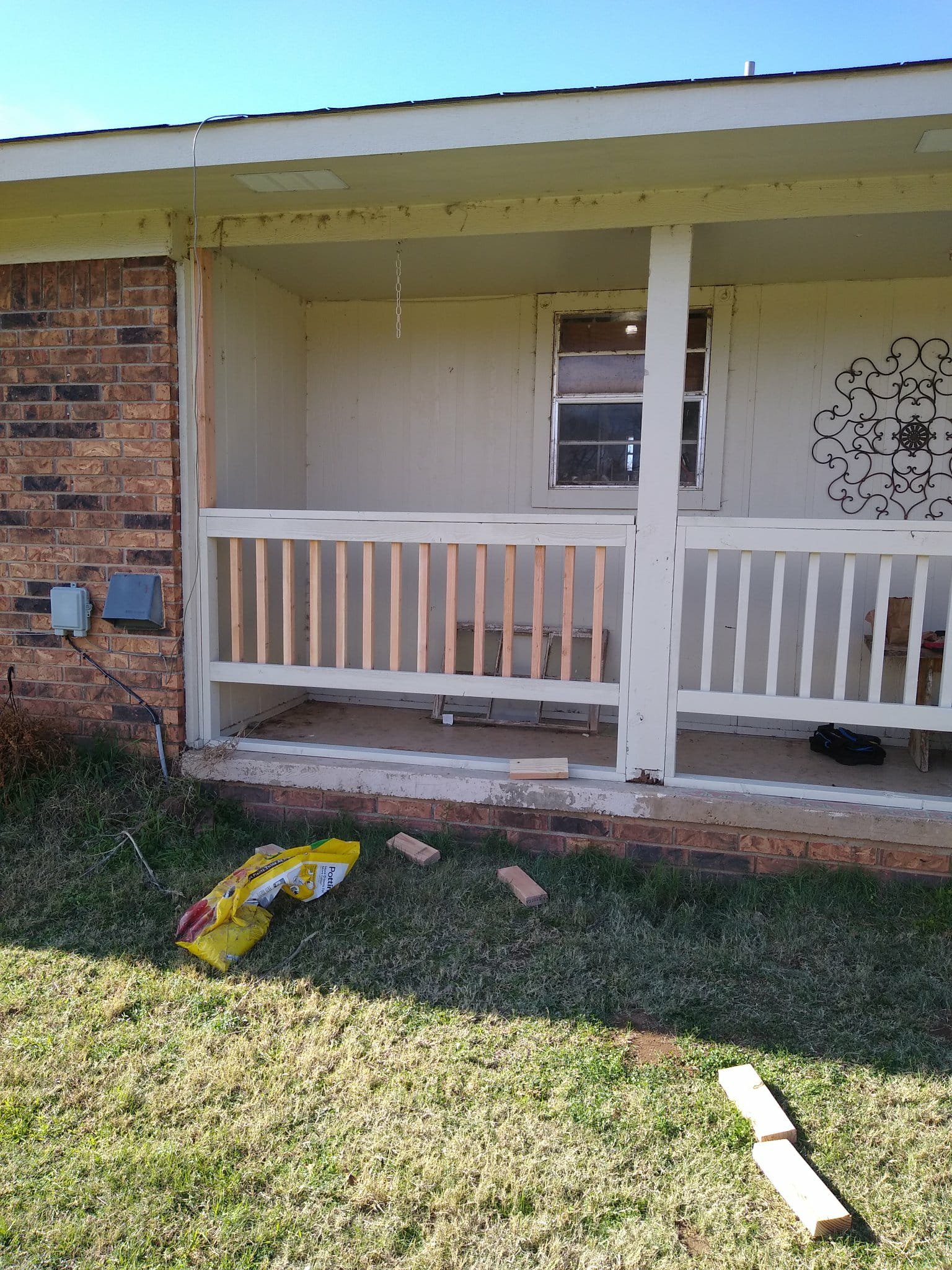 Front porch of a house under construction with white railing, window, and decorative metal wall art, with construction materials and tools on the ground.