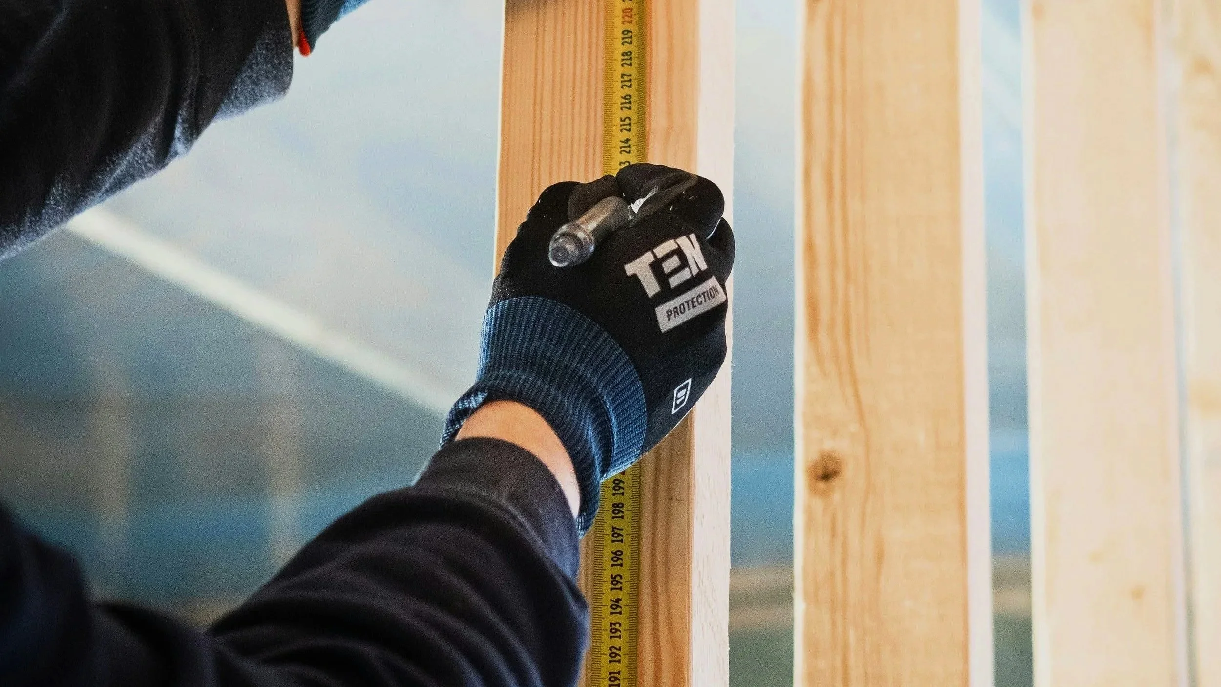Person wearing a black glove marked 'TEX PROTECTION' measuring wood with a yellow tape measure.