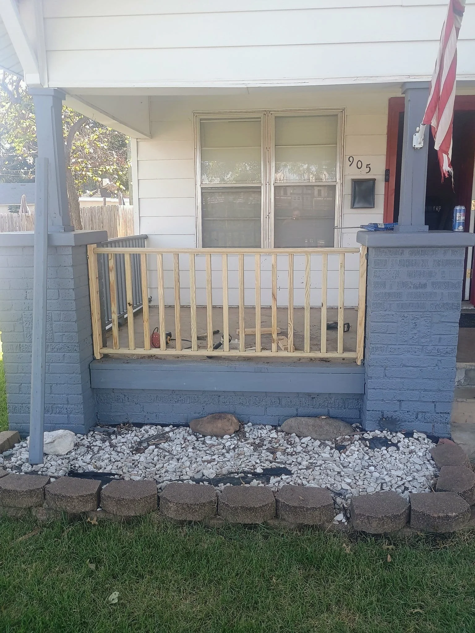 Front porch of a house with a newly built wooden railing, with a small garden bed with white rocks and large stones, and the house has white siding and a window with blinds, a mailbox, and a flag on a pole.