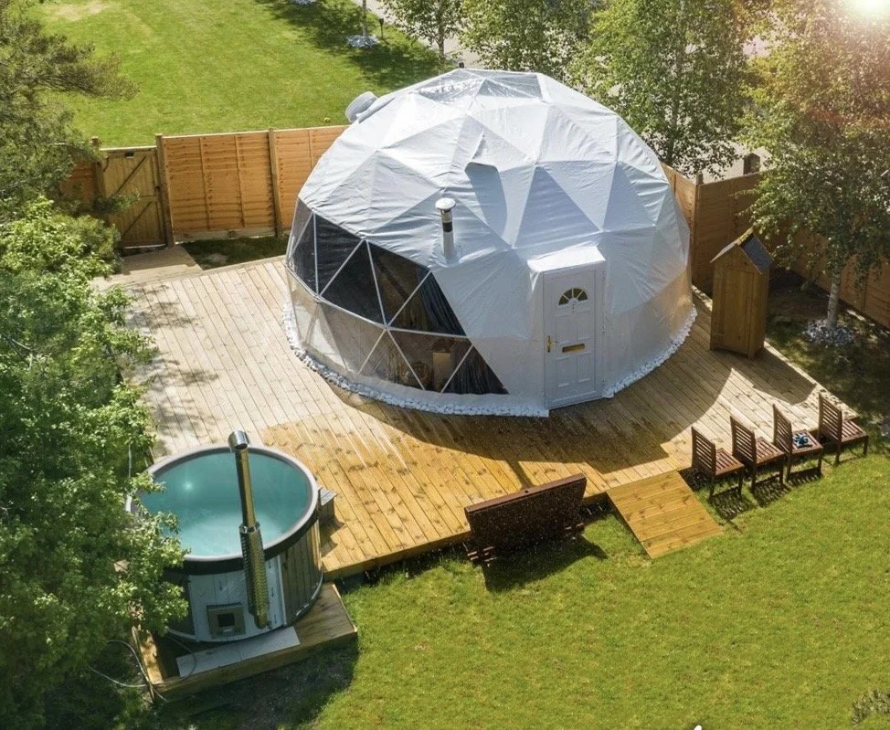 An outdoor backyard scene with a geodesic dome structure, a hot tub, a wooden deck, multiple chairs, and a picnic table, enclosed by a wooden fence and surrounded by trees.