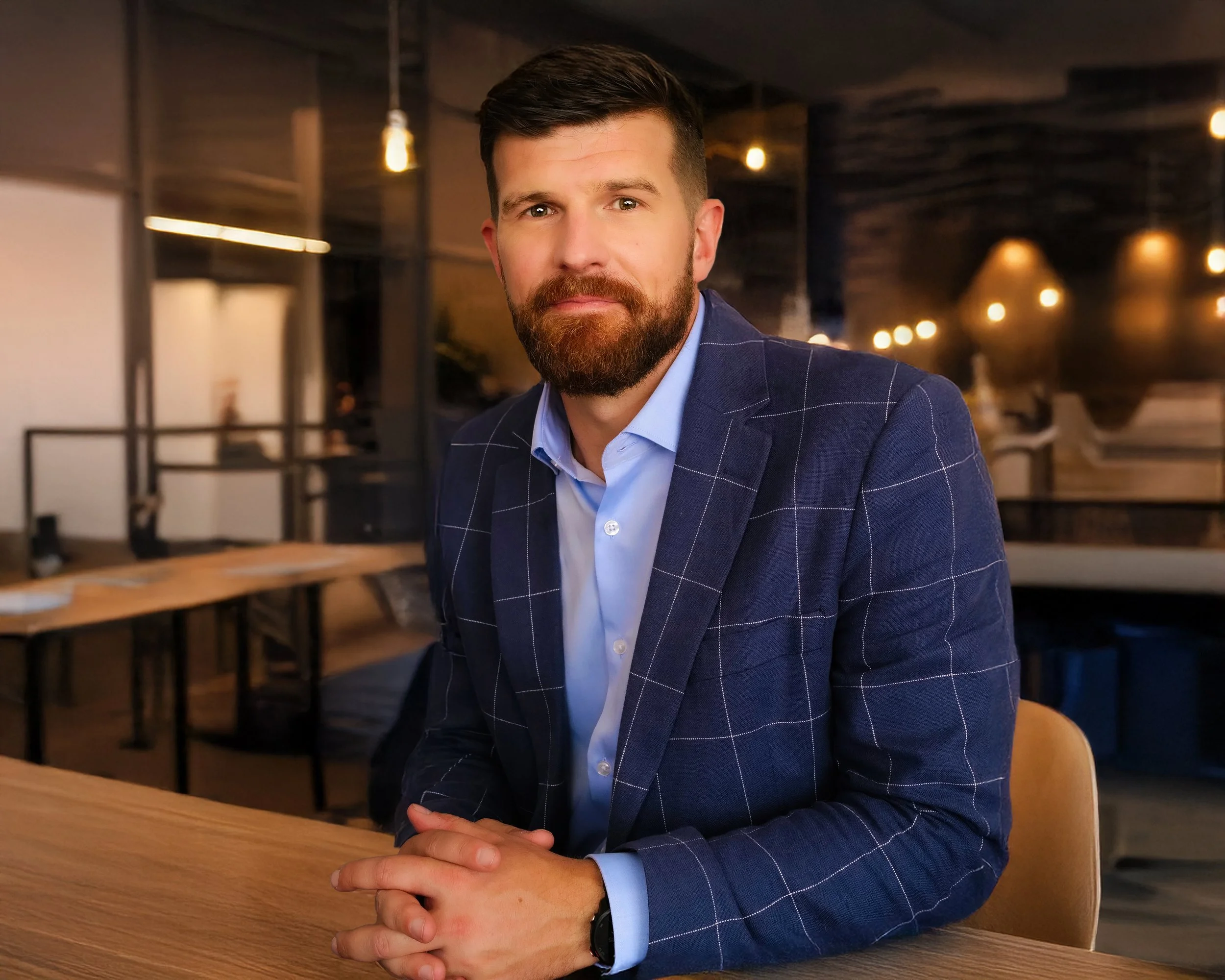 A learned lawyer with a beard and short hair, wearing a blue checked blazer and light blue shirt, sits awaiting instructions at a wooden table in a dimly lit modern restaurant or cafe.