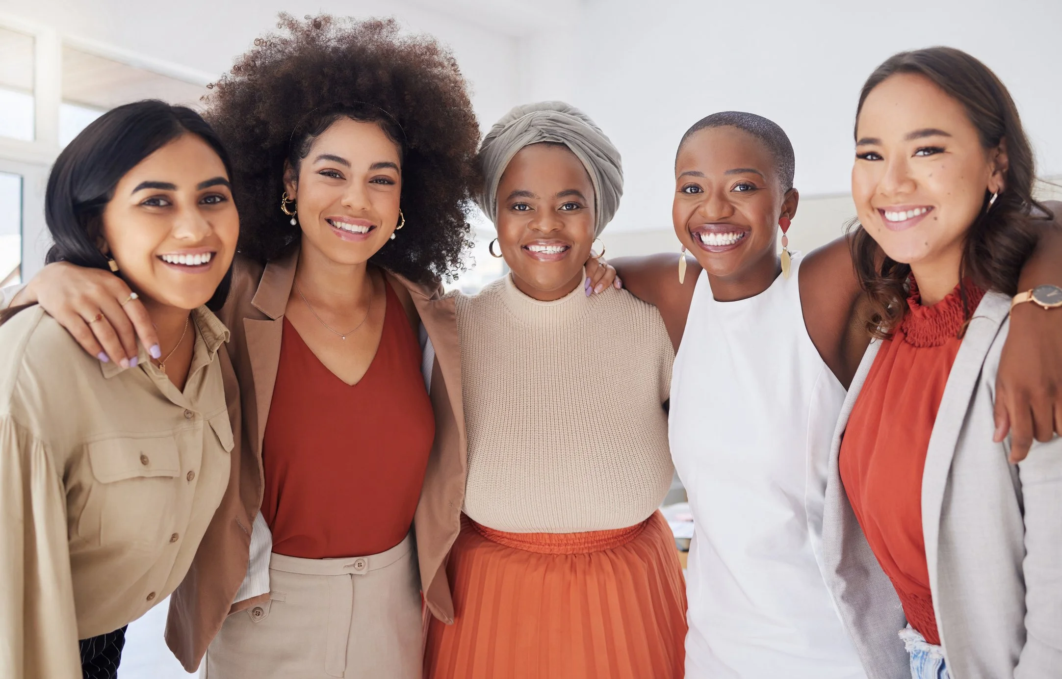 Five diverse women standing close together, smiling, with arms around each other in a bright, modern room.