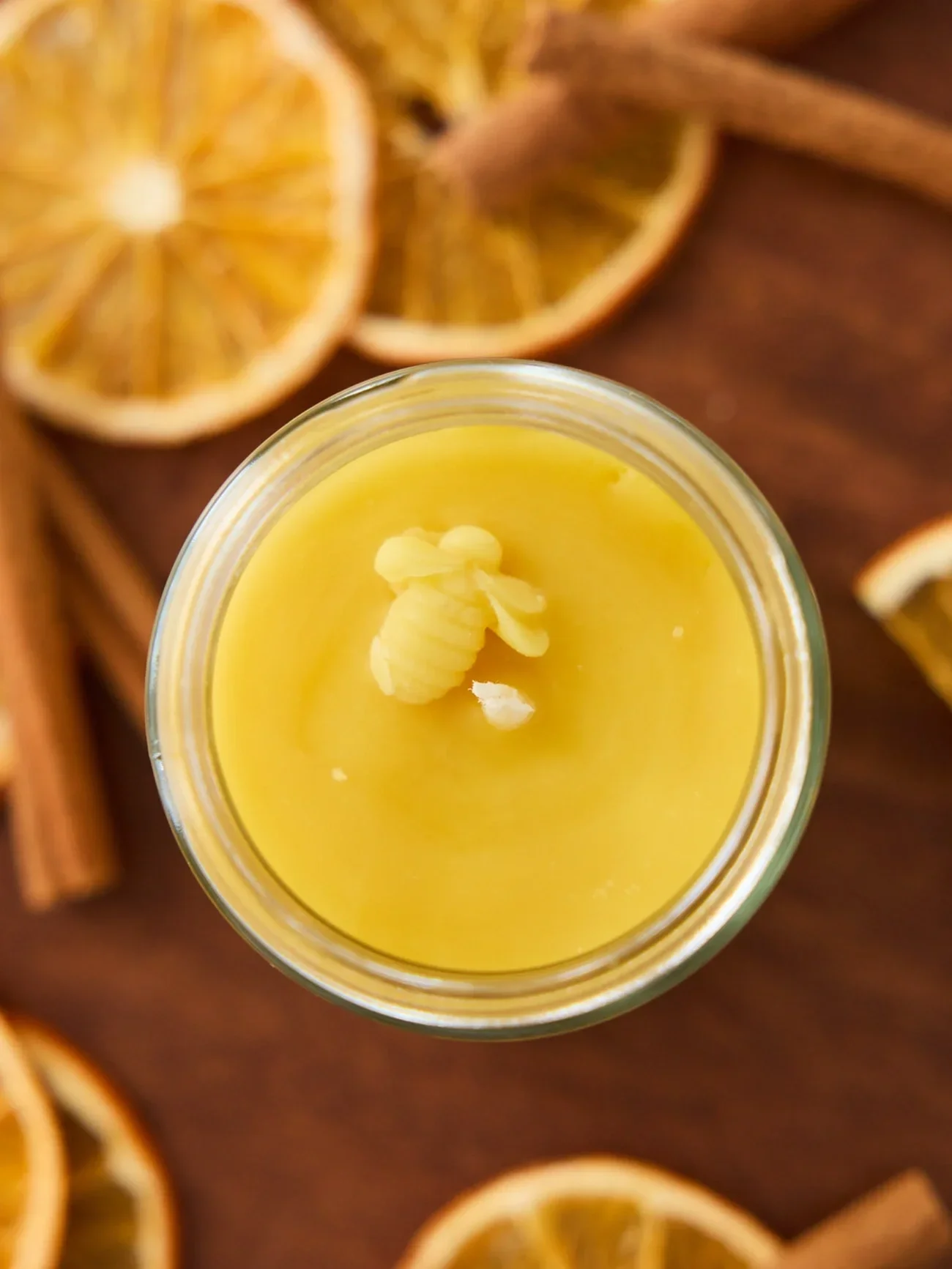 Close-up of a yellow beeswax candle with a white wick placed on a wooden surface surrounded by dried orange slices and cinnamon sticks.