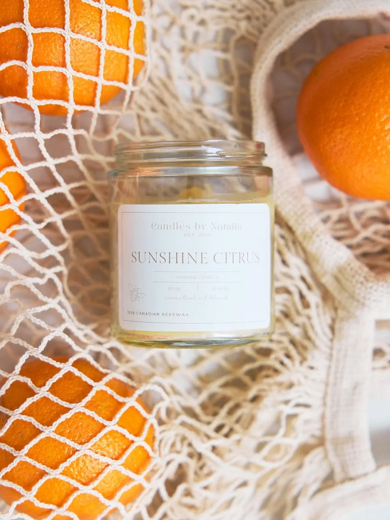A beeswax candle labeled 'Sunshine Citrus' surrounded by oranges in netted and fabric bags.
