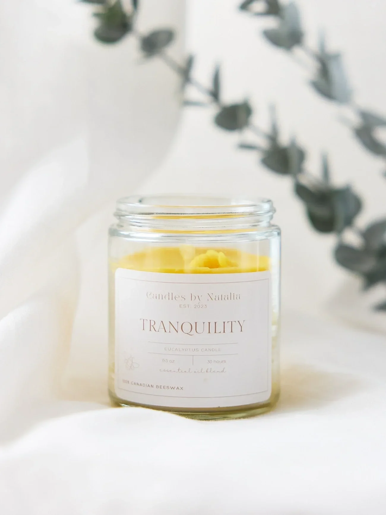 'Tranquility' candle on a white background and a blurred eucalyptus plant in the background.