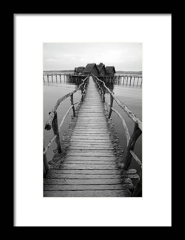 Black and white photo of a weathered wooden pier leading to a house on stilts over water, with calm water surroundings.
