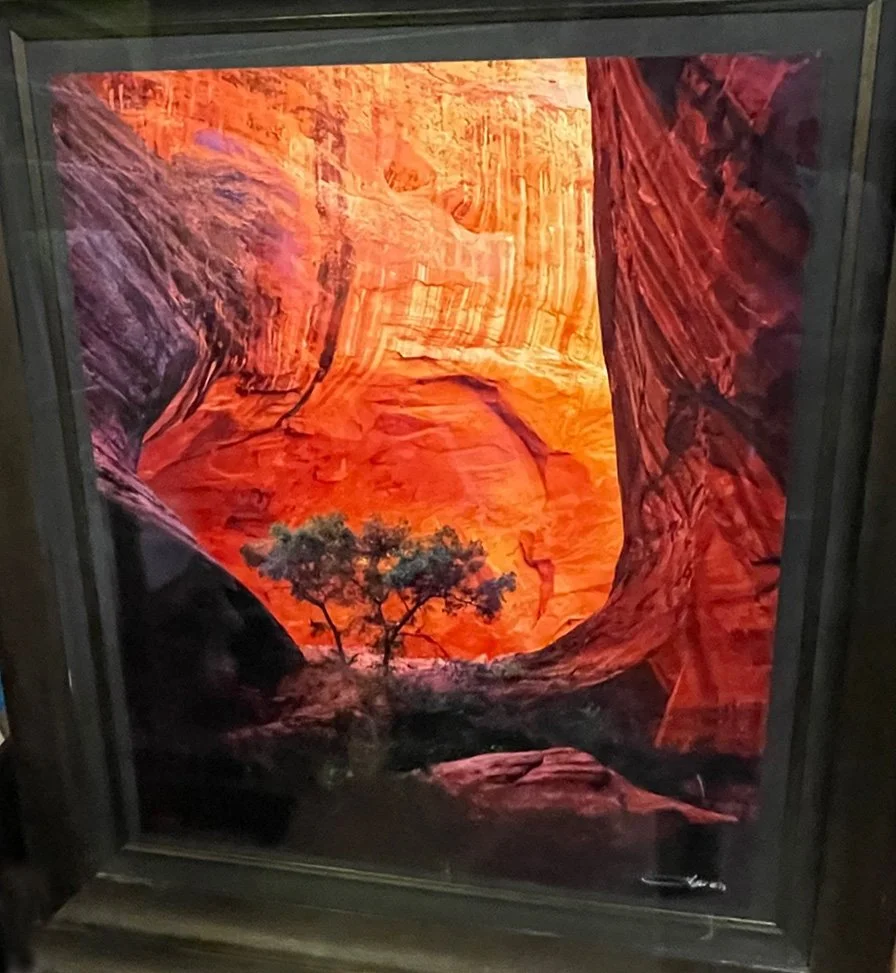 Photograph of a framed painting depicting a canyon landscape with tall orange and red rock formations, a sky with warm hues, and a few trees in the foreground.
