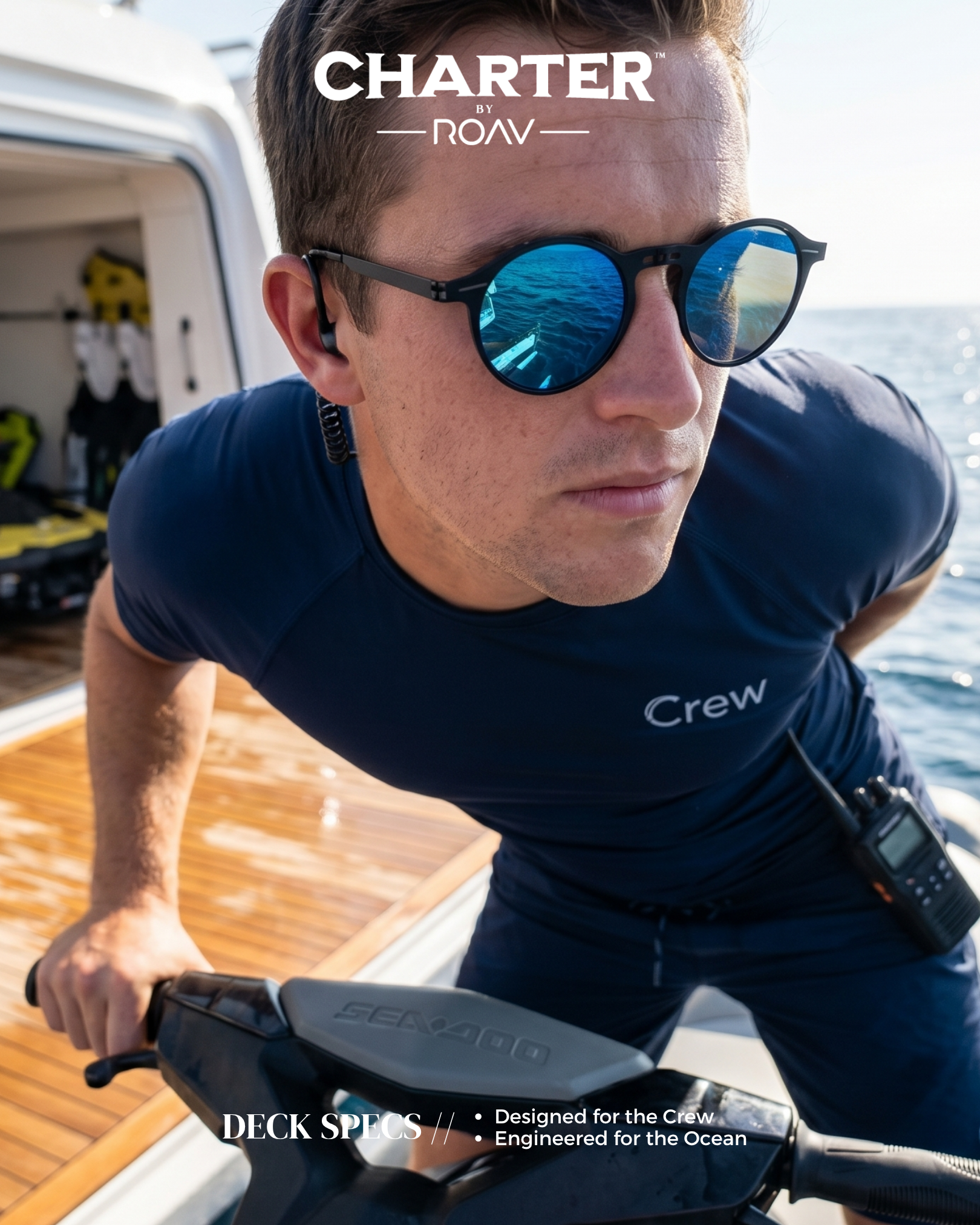 A man wearing sunglasses, a navy blue crew t-shirt, and holding a handlebar, on a boat with a wooden deck during daytime. The reflection in the sunglasses shows the ocean and boat equipment.