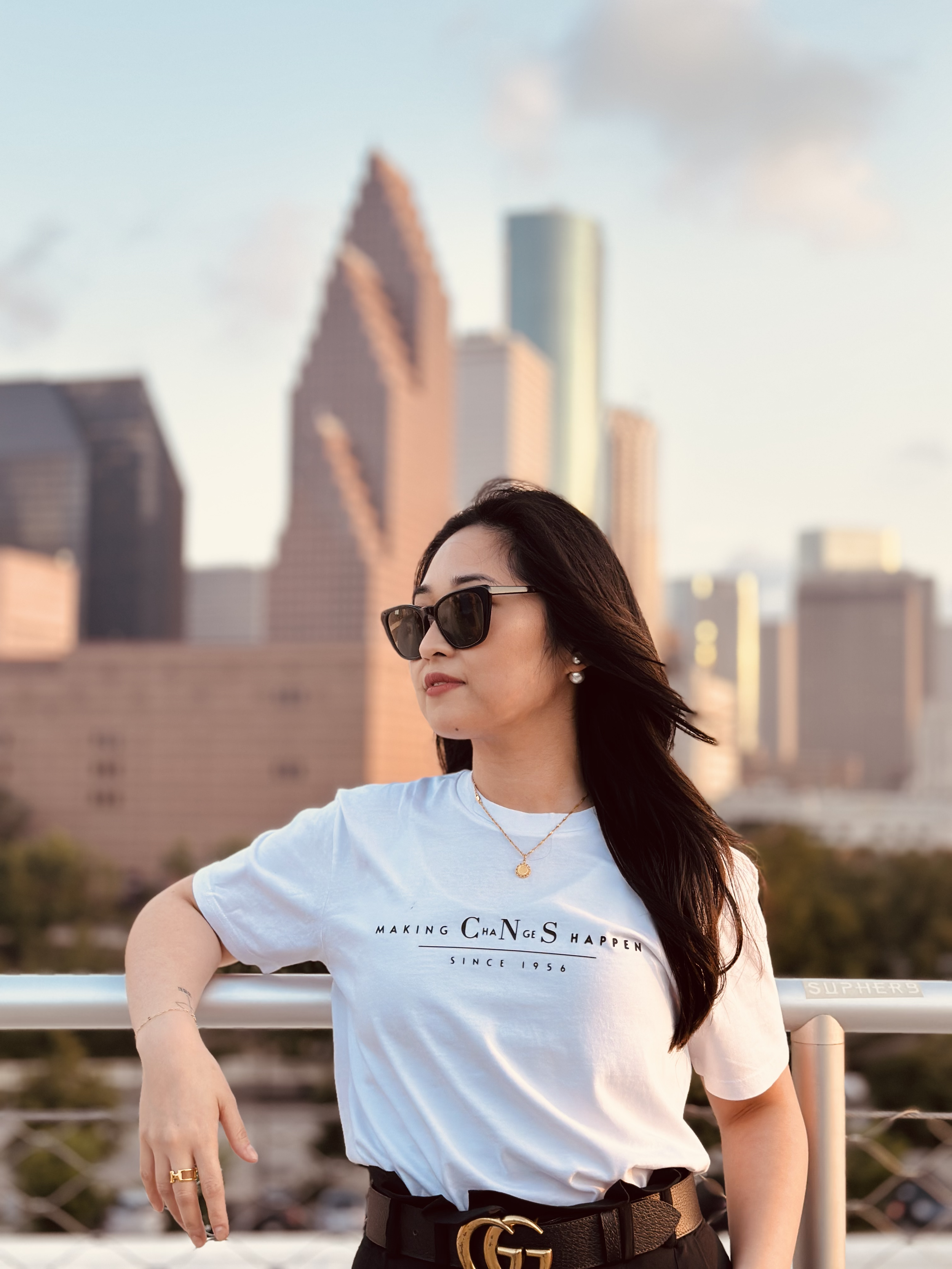 Woman with sunglasses and jewelry standing on a rooftop with city skyscrapers in the background during sunset.