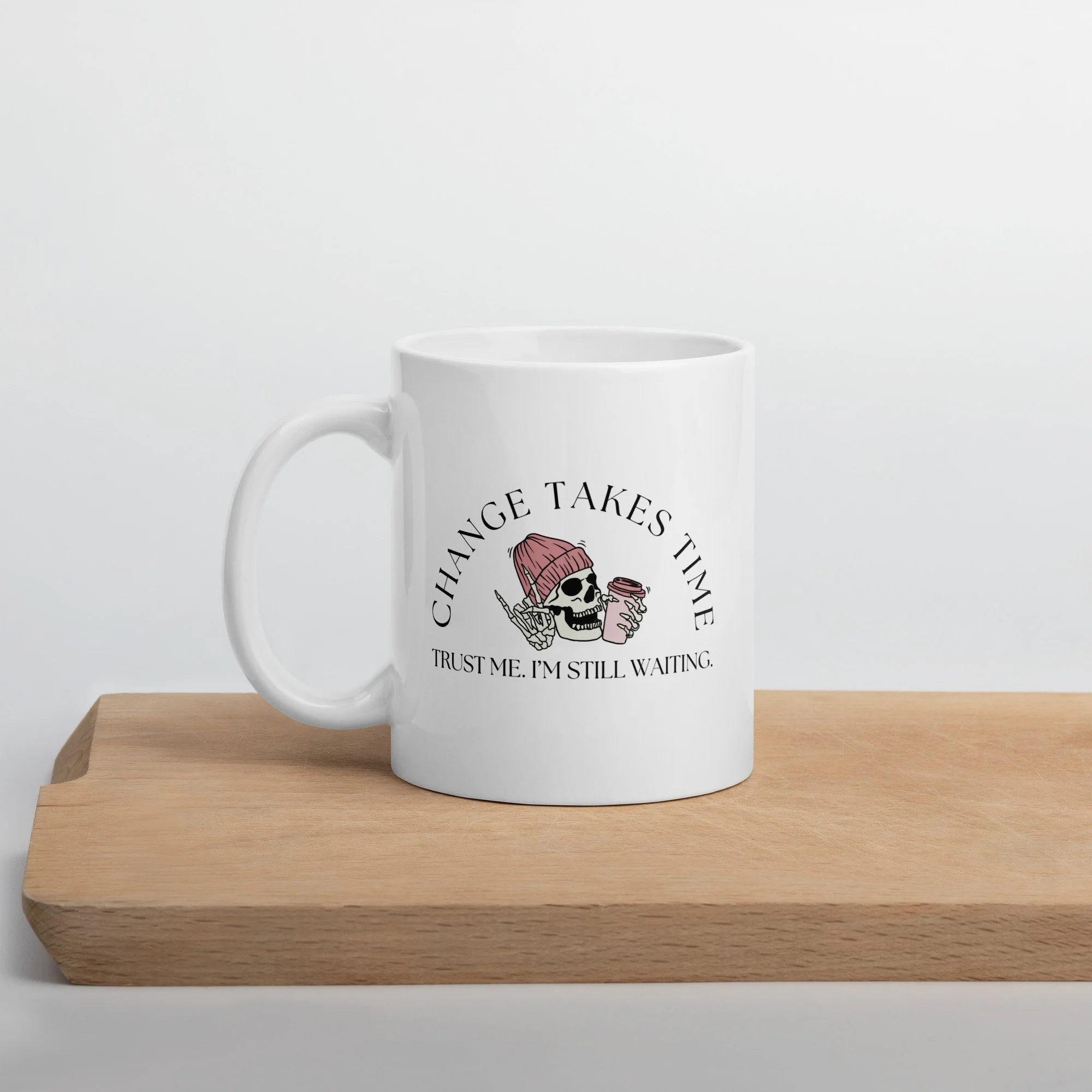 A white ceramic mug on a wooden board, featuring a design of a skull wearing a pink beanie holding a pink coffee cup with the text "Change Takes Time. Trust me, I’m still waiting."
