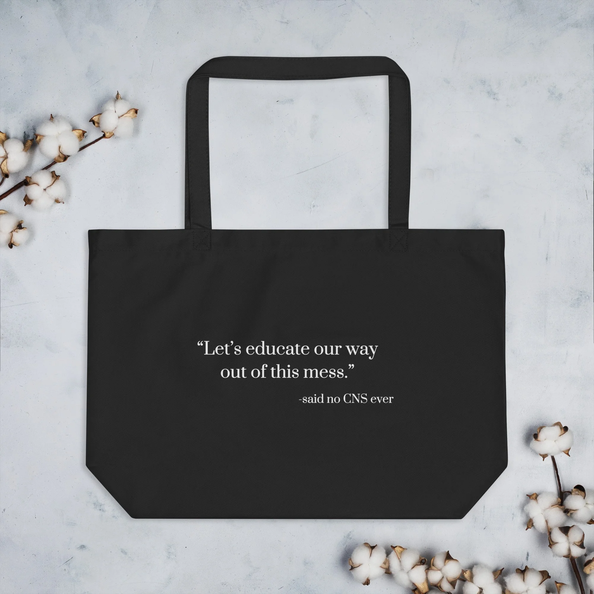 Black tote bag with white text that says "Let's educate our way out of this mess." -said no CNS ever, surrounded by cotton flowers on a light gray surface.