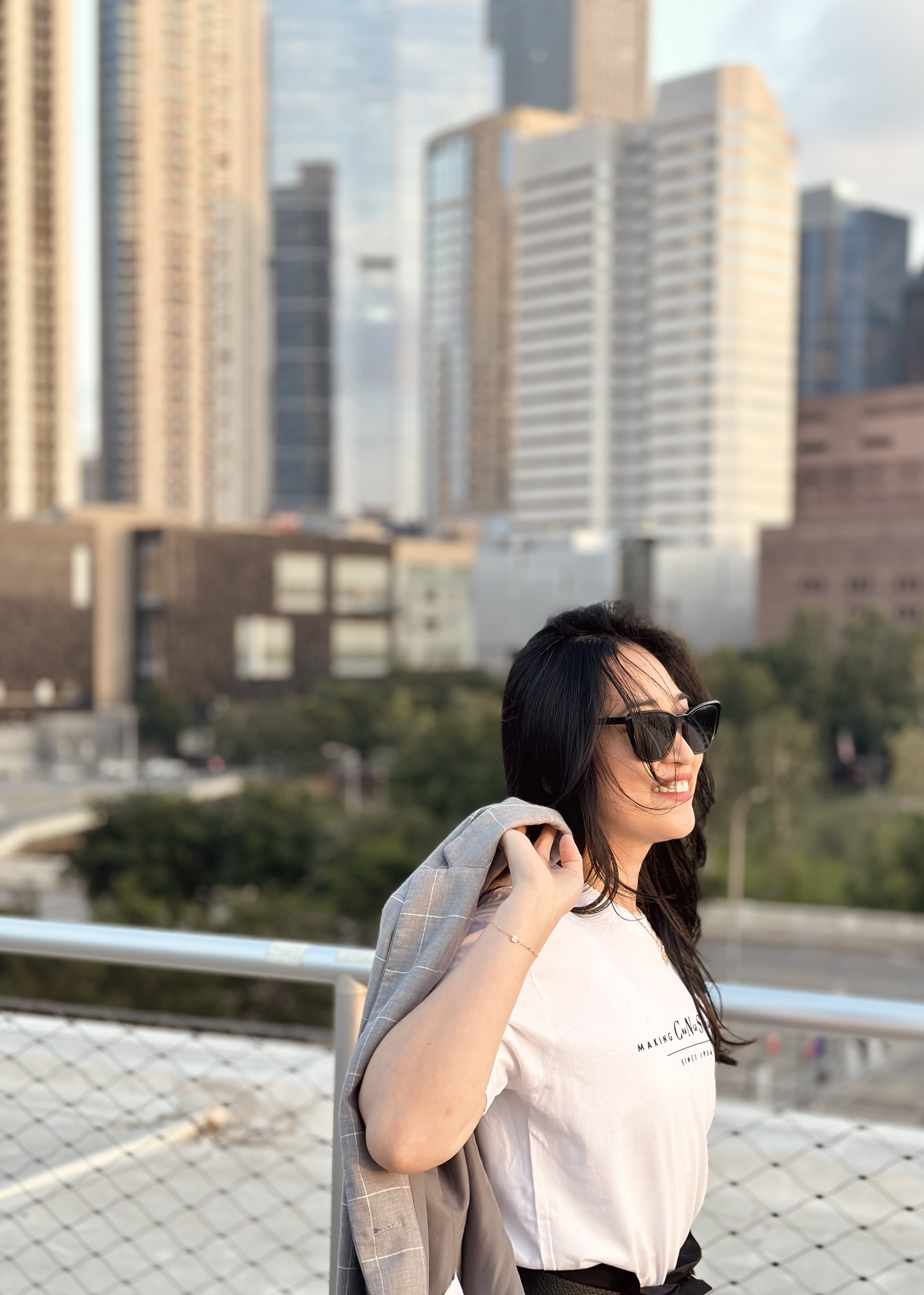 A woman with dark hair wearing sunglasses, a white t-shirt, and a blazer over her shoulder, smiling outdoors with a city skyline in the background.