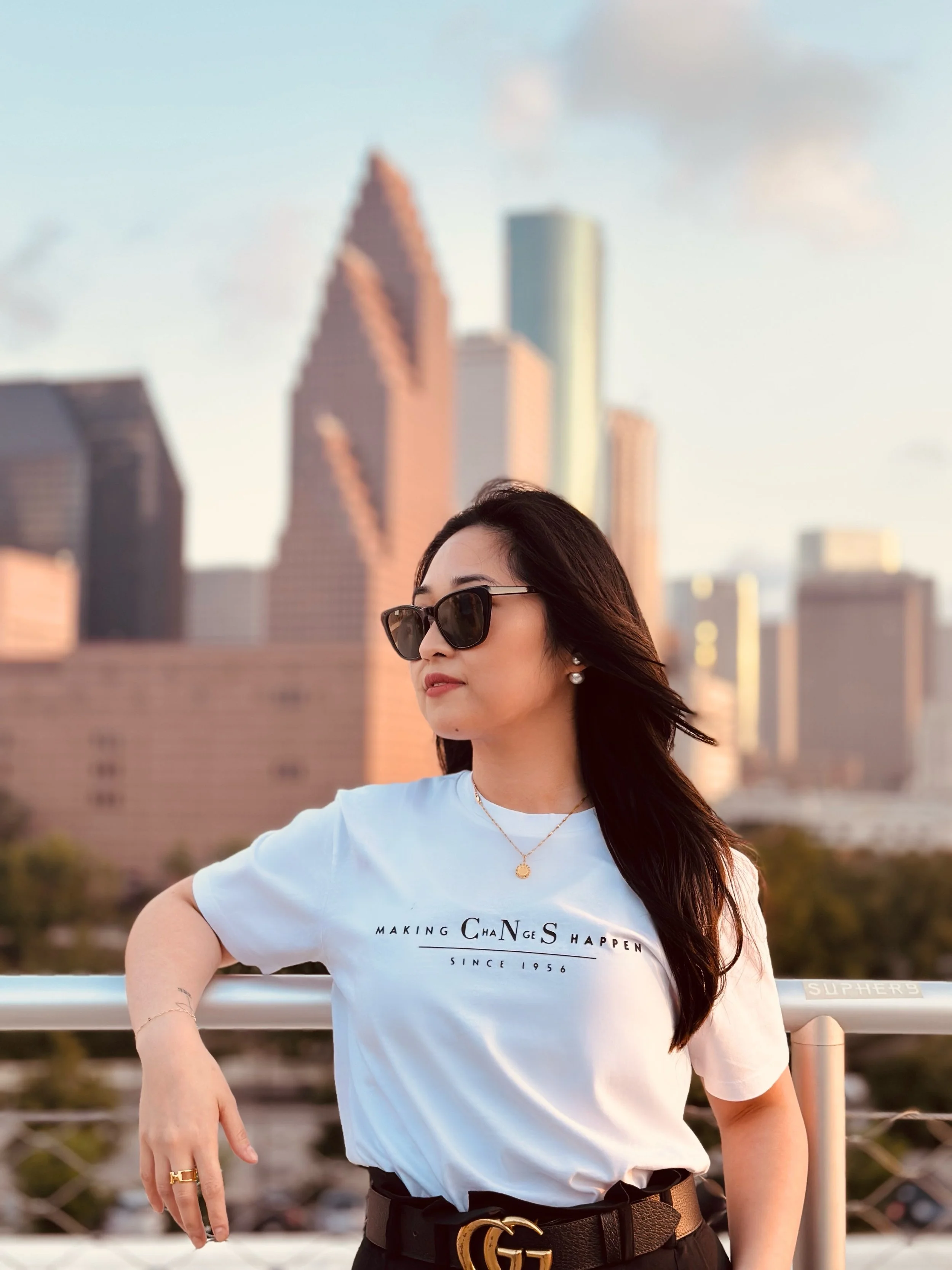 A woman with long dark hair wearing sunglasses and a white T-shirt with text, standing outdoors with a city skyline in the background during daytime.