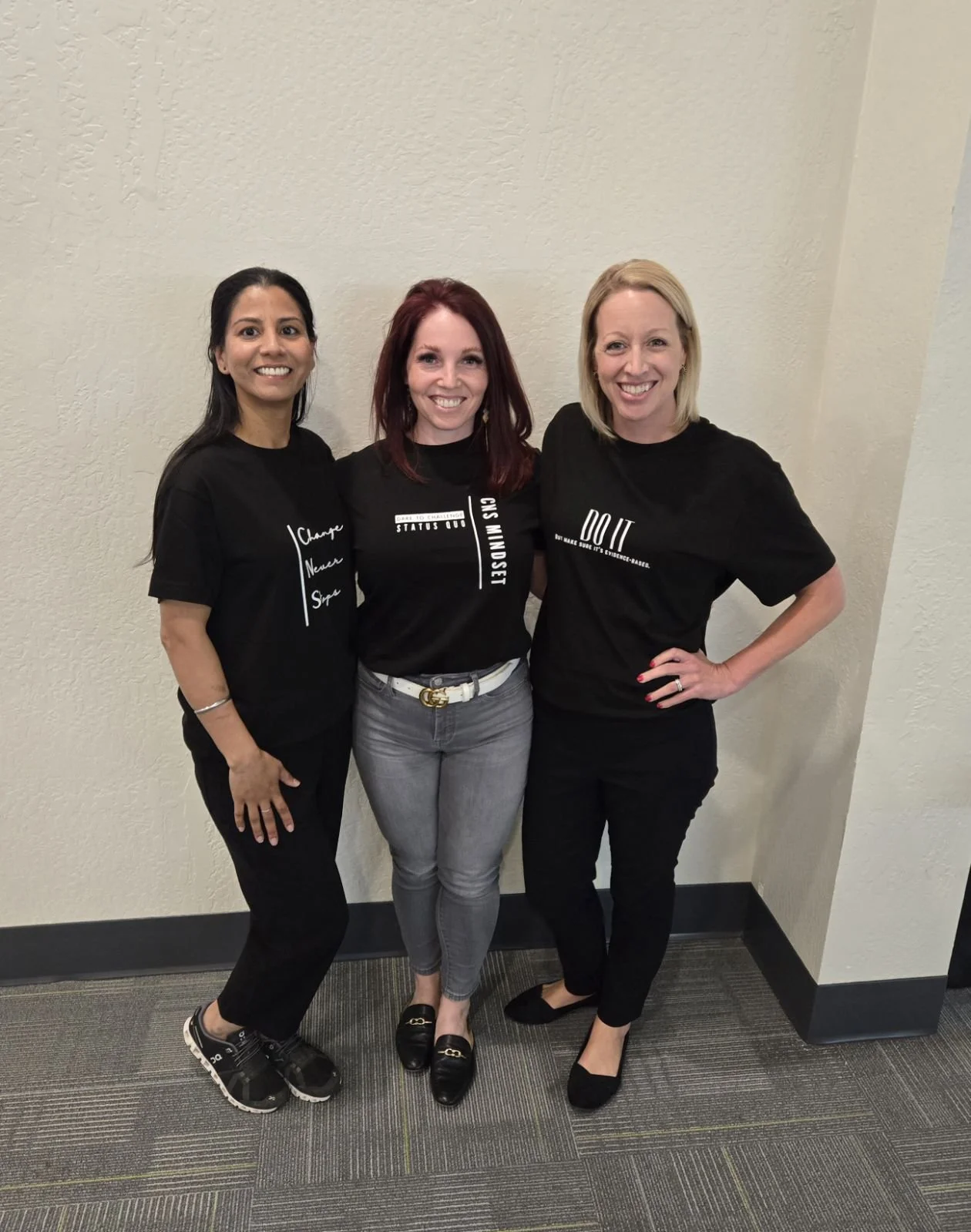Three women standing together indoors against a beige wall, smiling, all wearing black T-shirts with motivational phrases.