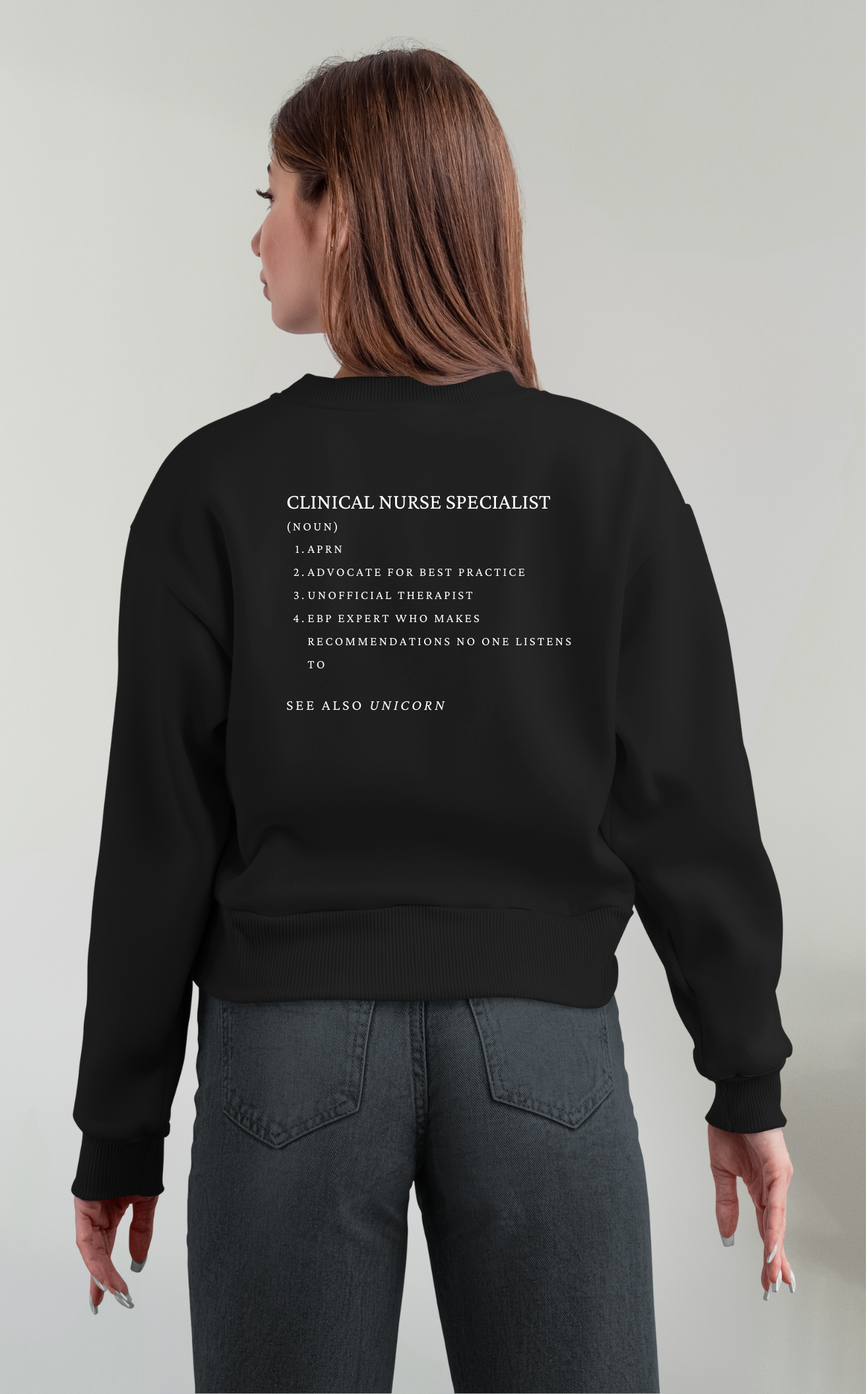 Back view of a person wearing a black sweatshirt with humorous text about a clinical nurse specialist, and gray jeans, standing against a light-colored background.