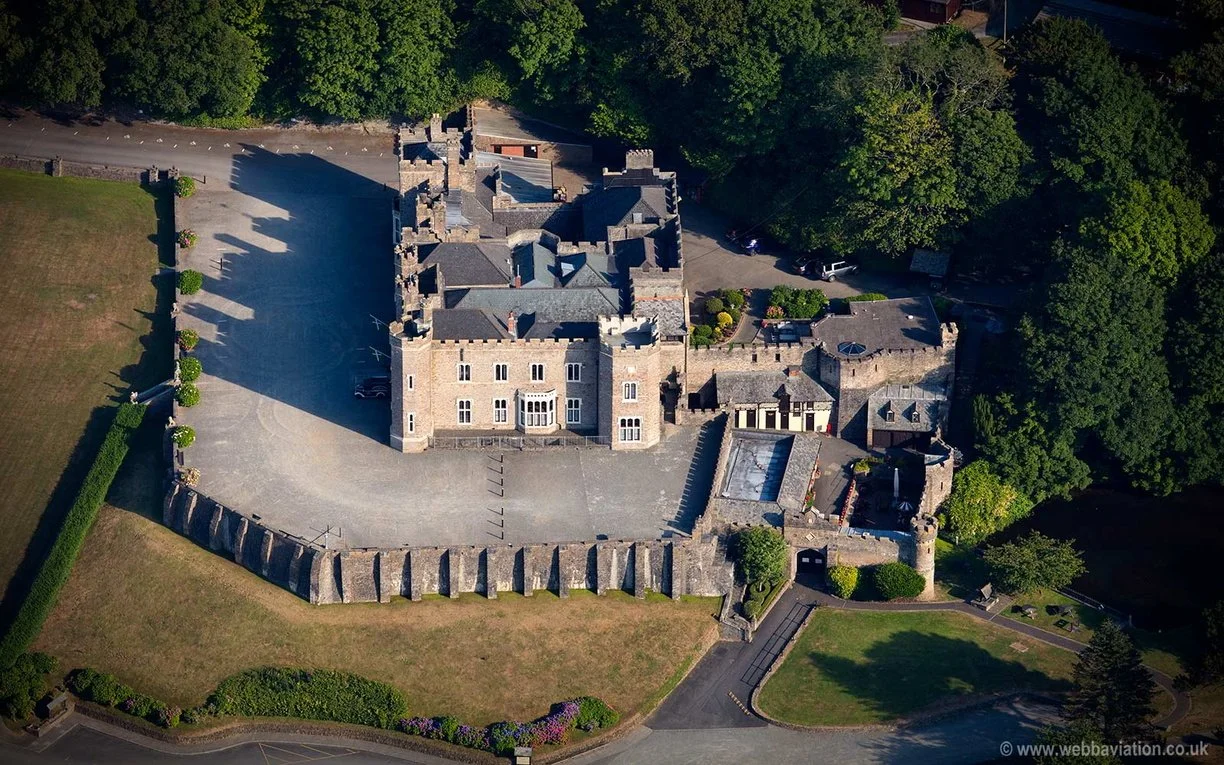 Aerial view of a castle with a large stone courtyard surrounded by trees and green gardens. The castle has multiple towers, battlements, and a main entrance with steps. There's a parking lot with marked spaces in front of the castle and a driveway leading to the main entrance.