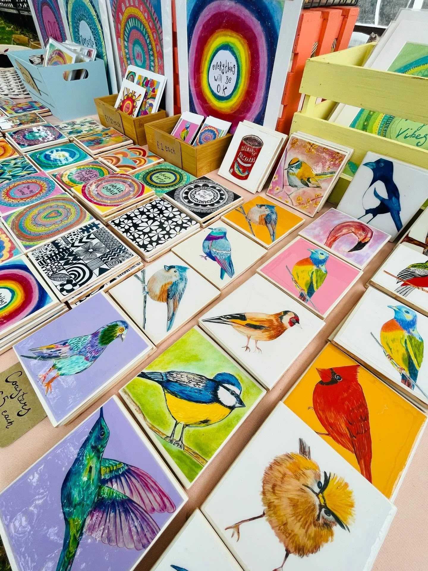A display of colorful bird and abstract art prints on a table, with some postcards and greeting cards featuring vibrant bird illustrations, patterned tiles, and circular rainbow designs.
