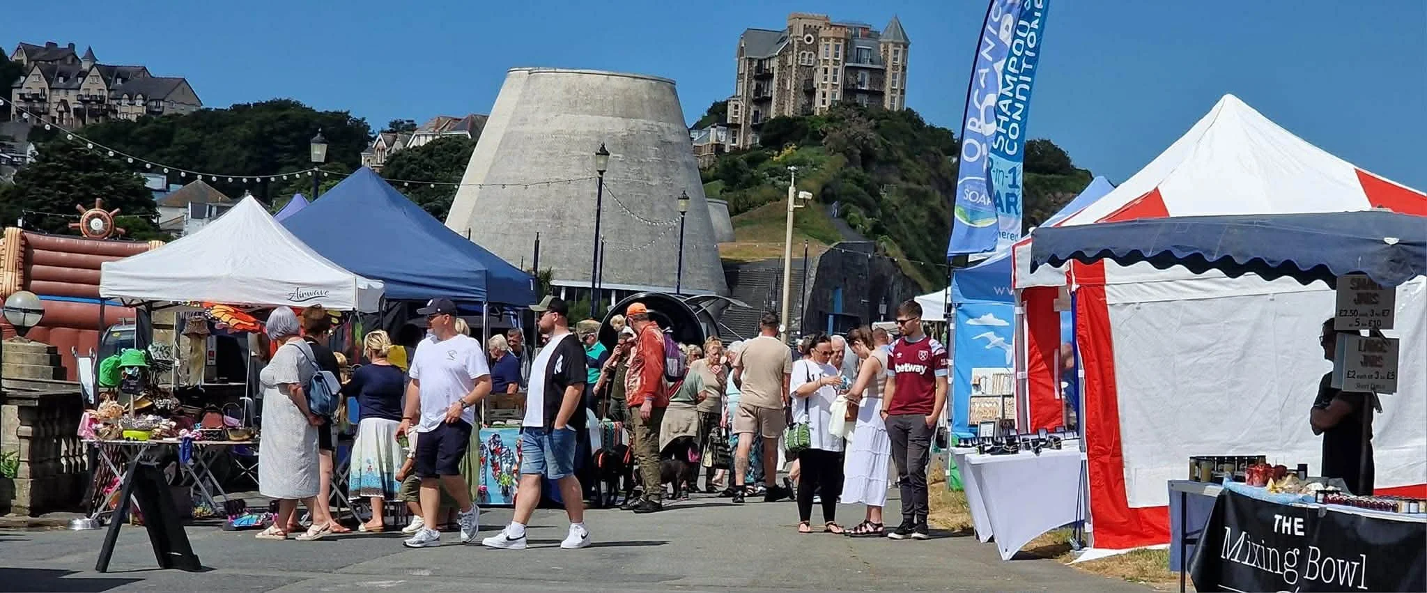 Outdoor market with stalls and tents, and people browsing and shopping near a large concrete structure and residential buildings on a hill in the background.