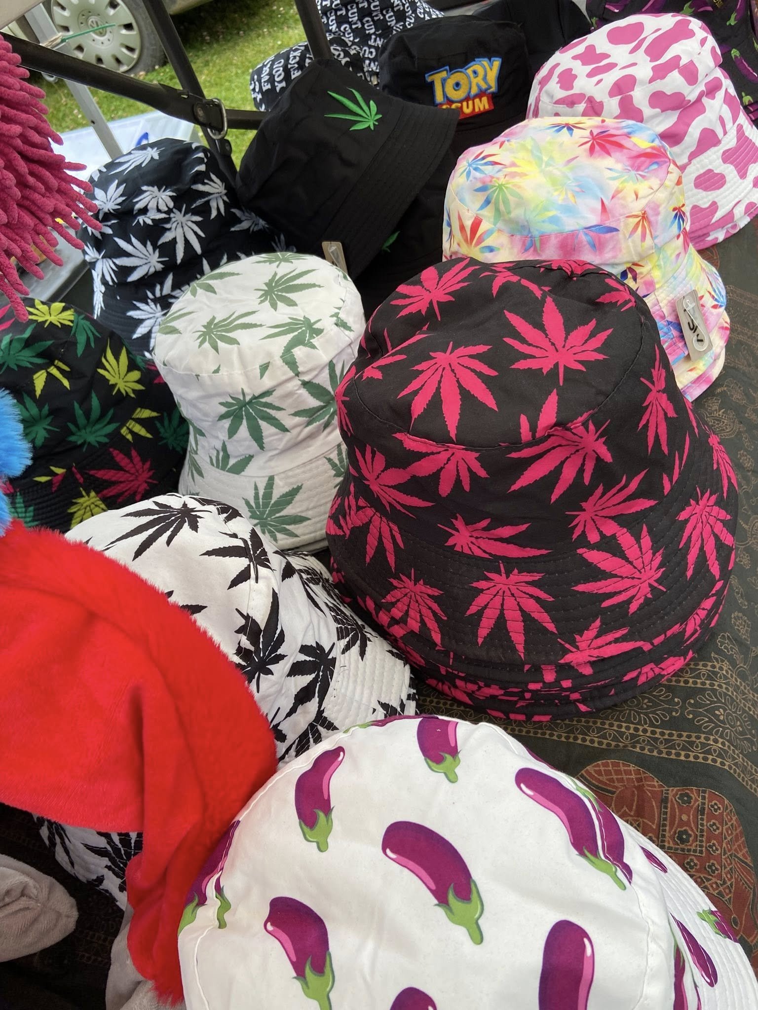 A display of hats with various colorful cannabis leaf patterns on a table at an outdoor market or event. The hats feature different colors and designs, including pink, green, black, and multicolored backgrounds.