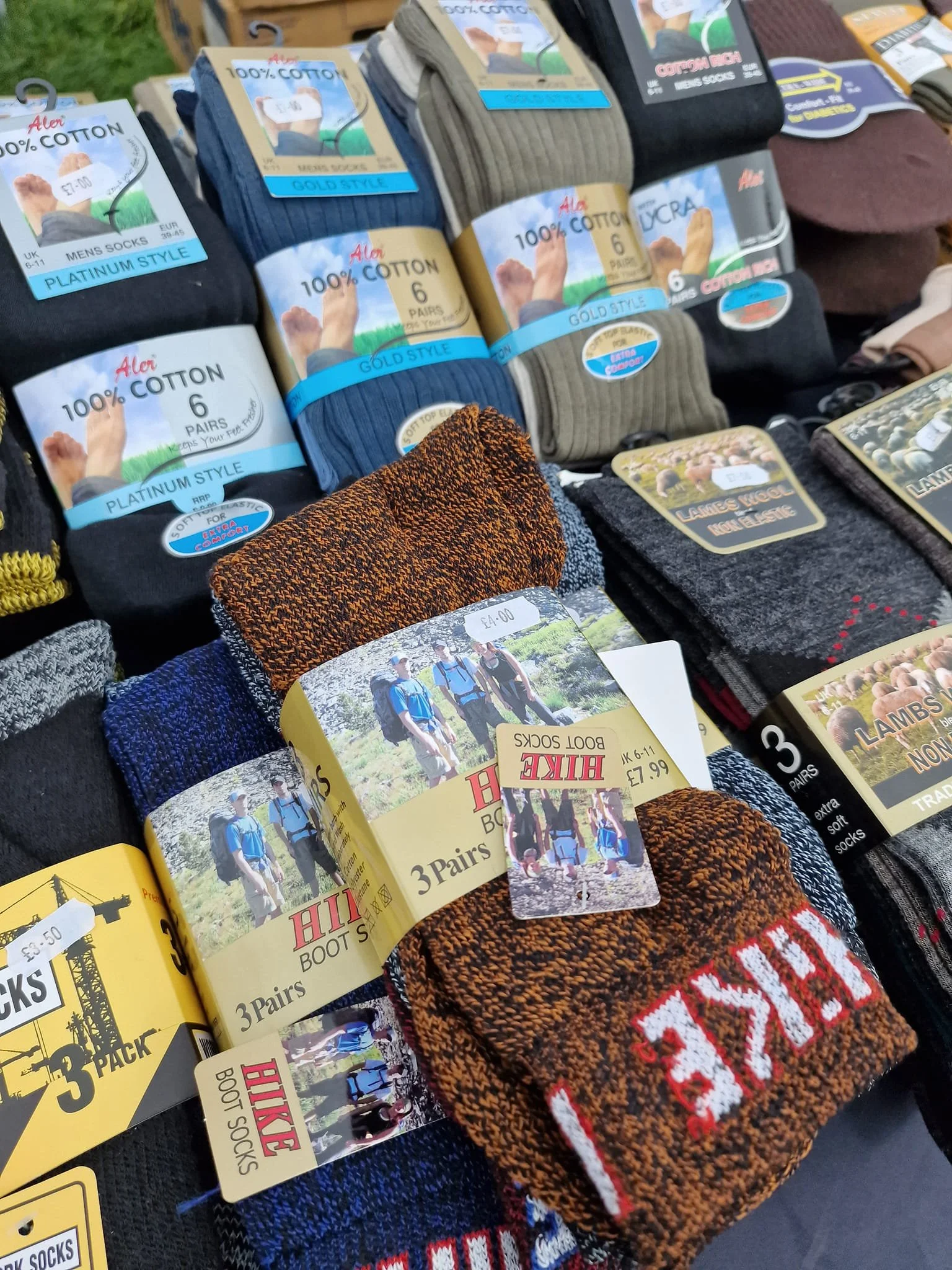 Display of men's socks in various colors and brands at a market stall, including cotton, fleece, and lambs wool socks, with price tags and packaging labels.